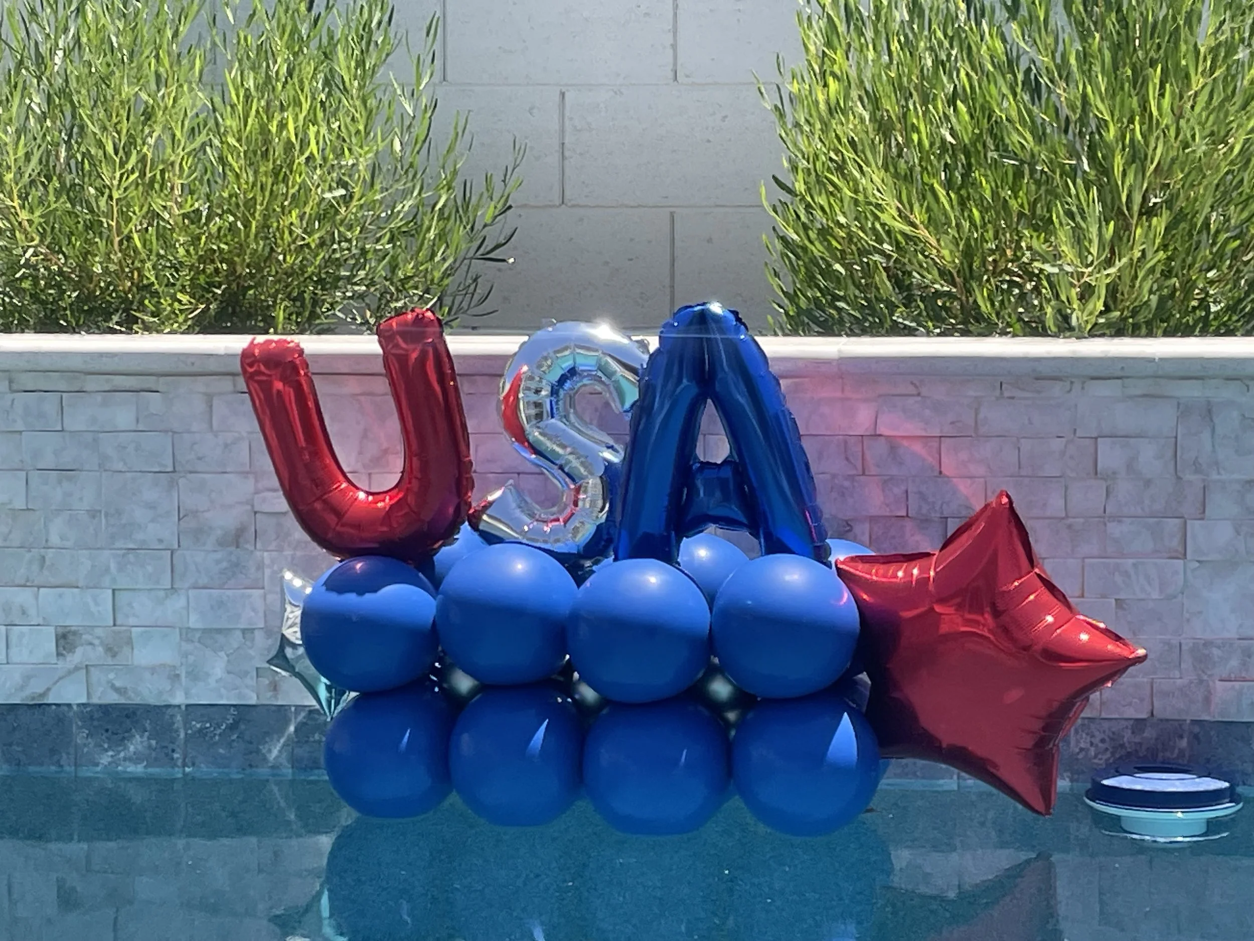 Colorful balloons arranged on the ground in front of a white brick wall, with green bushes and a gray concrete wall behind.