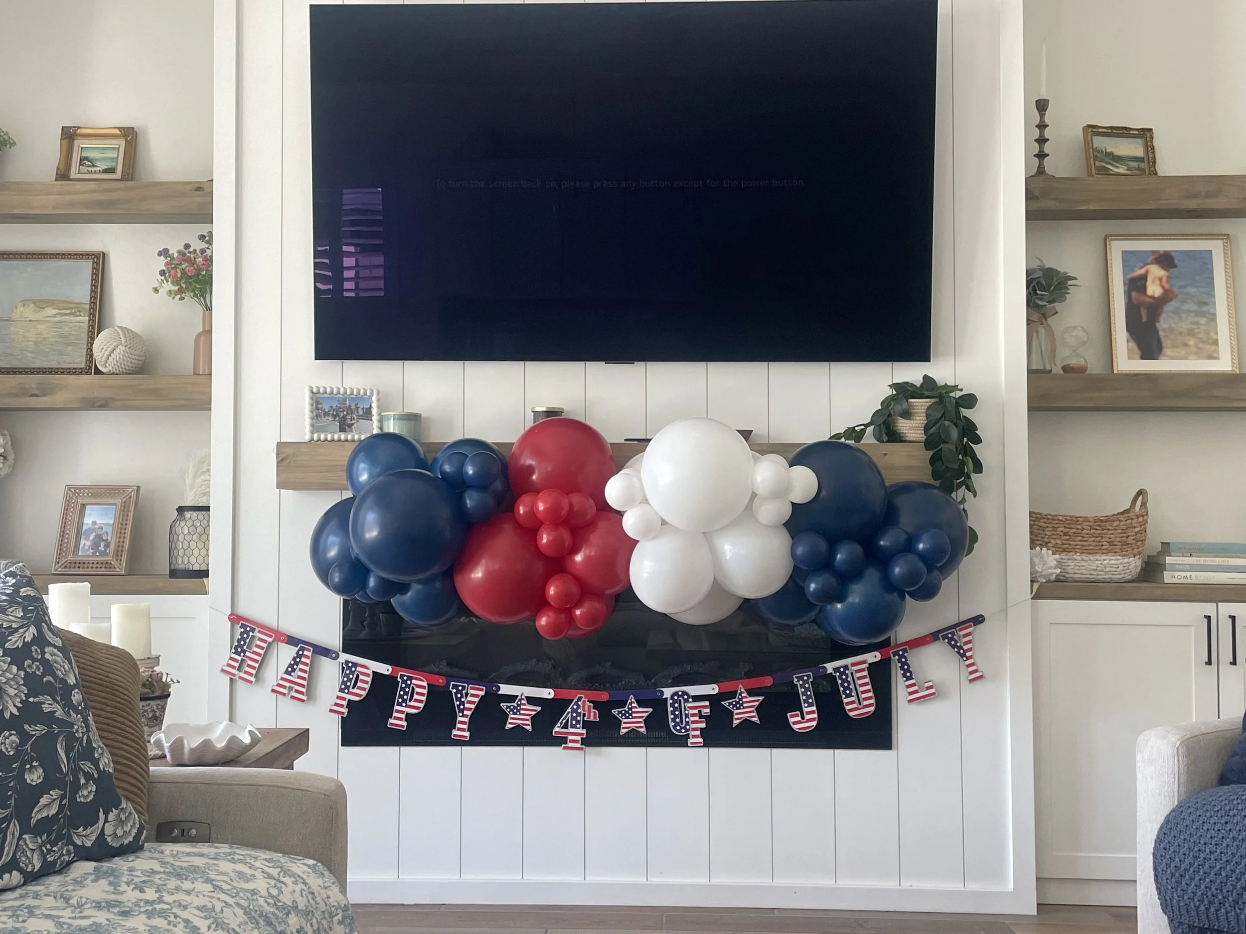 A living room decorated for a patriotic celebration with an American flag-themed banner, red, white, and blue balloons, and a mantel with framed pictures and decorative items.
