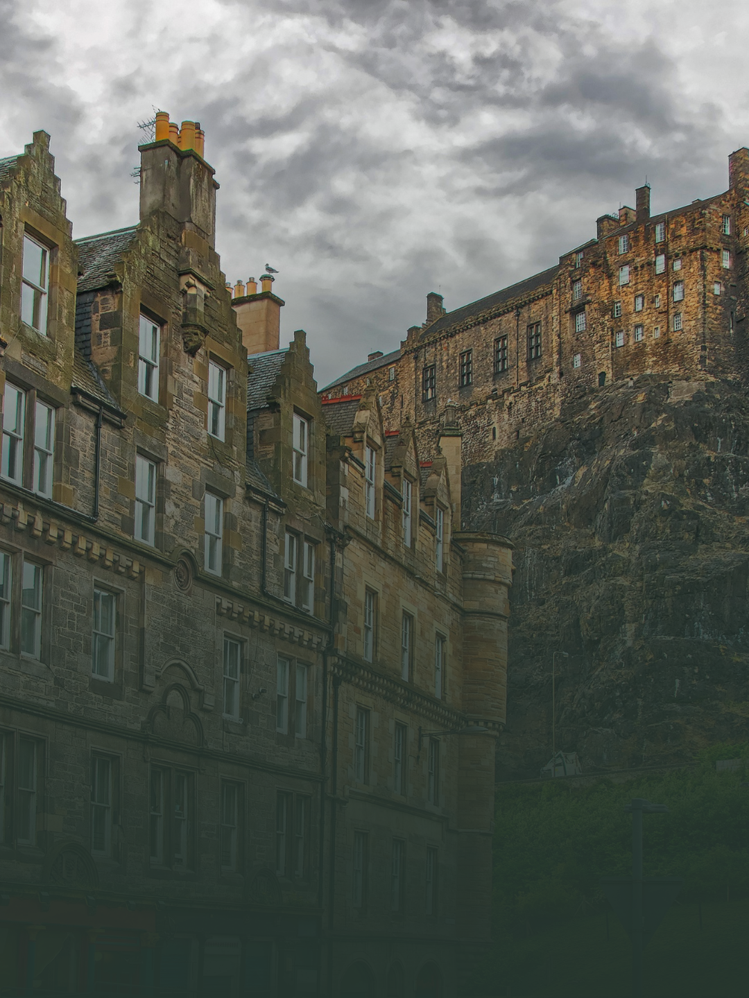 Historic stone buildings with multiple chimneys on a rocky hillside under a cloudy sky.