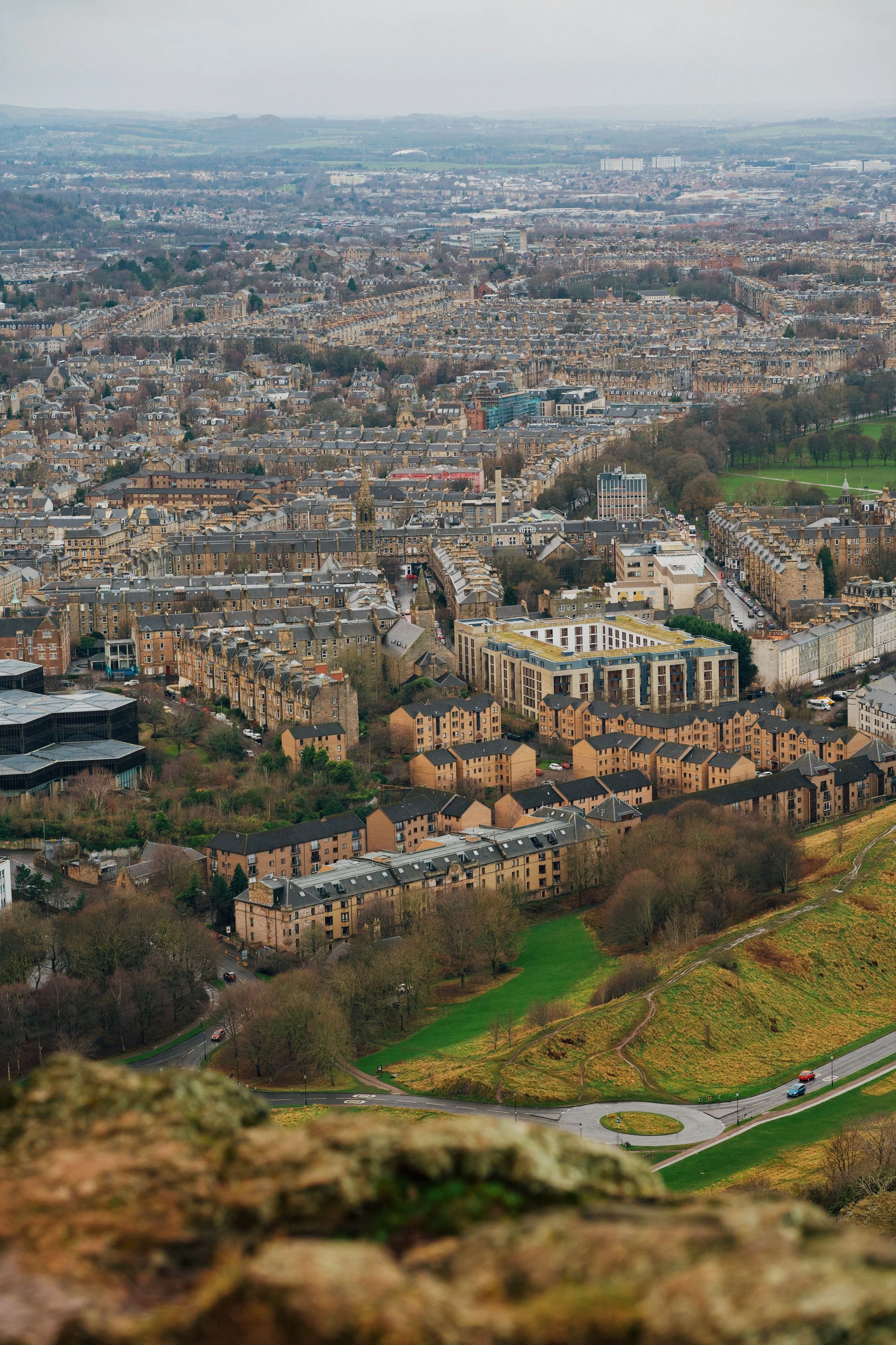 A panoramic view of a cityscape with numerous residential buildings, green parks, and winding roads under an overcast sky, as seen from a high vantage point.
