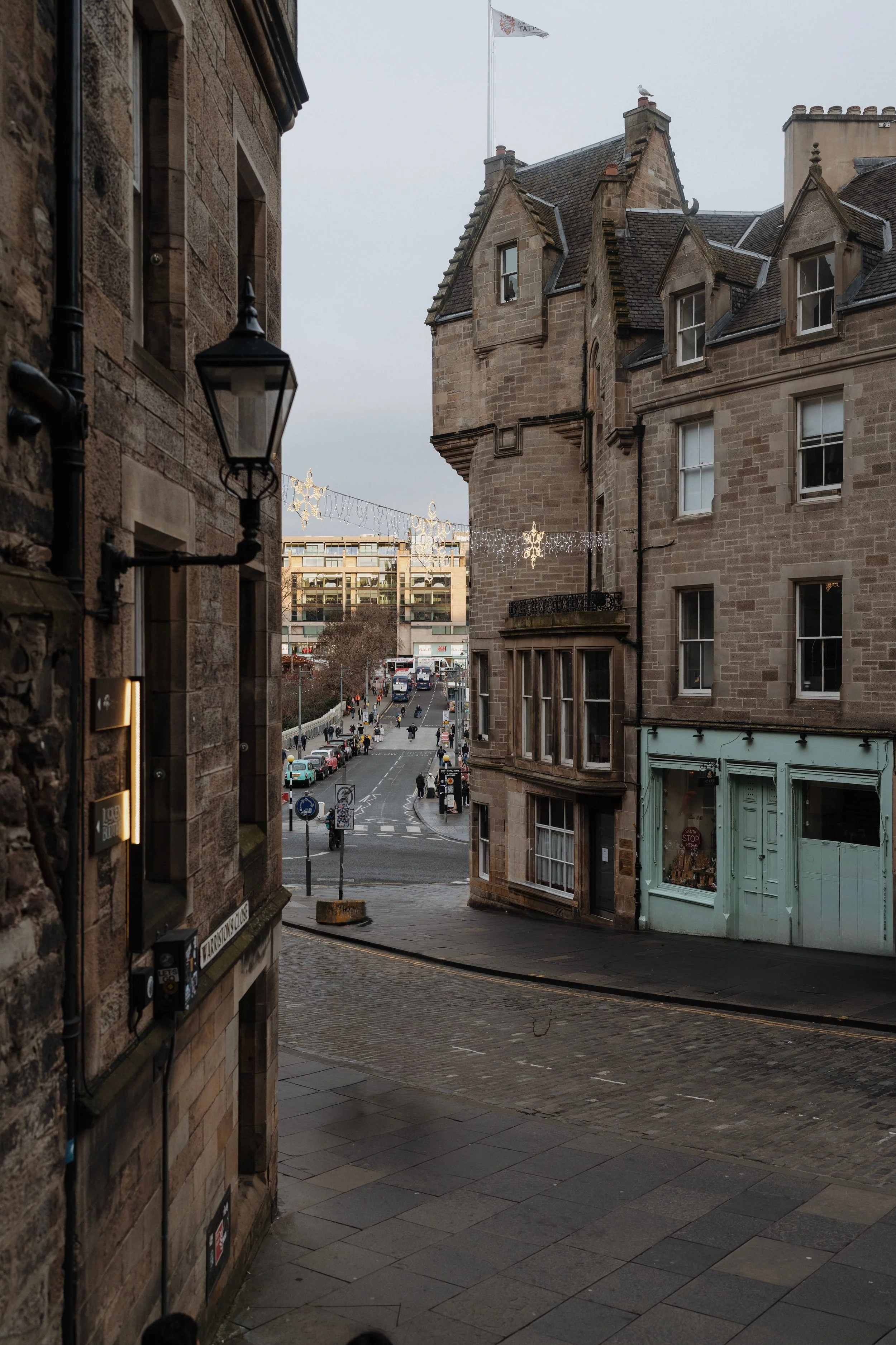 A street scene viewed from a narrow alleyway with historic stone buildings on either side, and holiday lights and a flag with a seagull on top of a building in the distance.