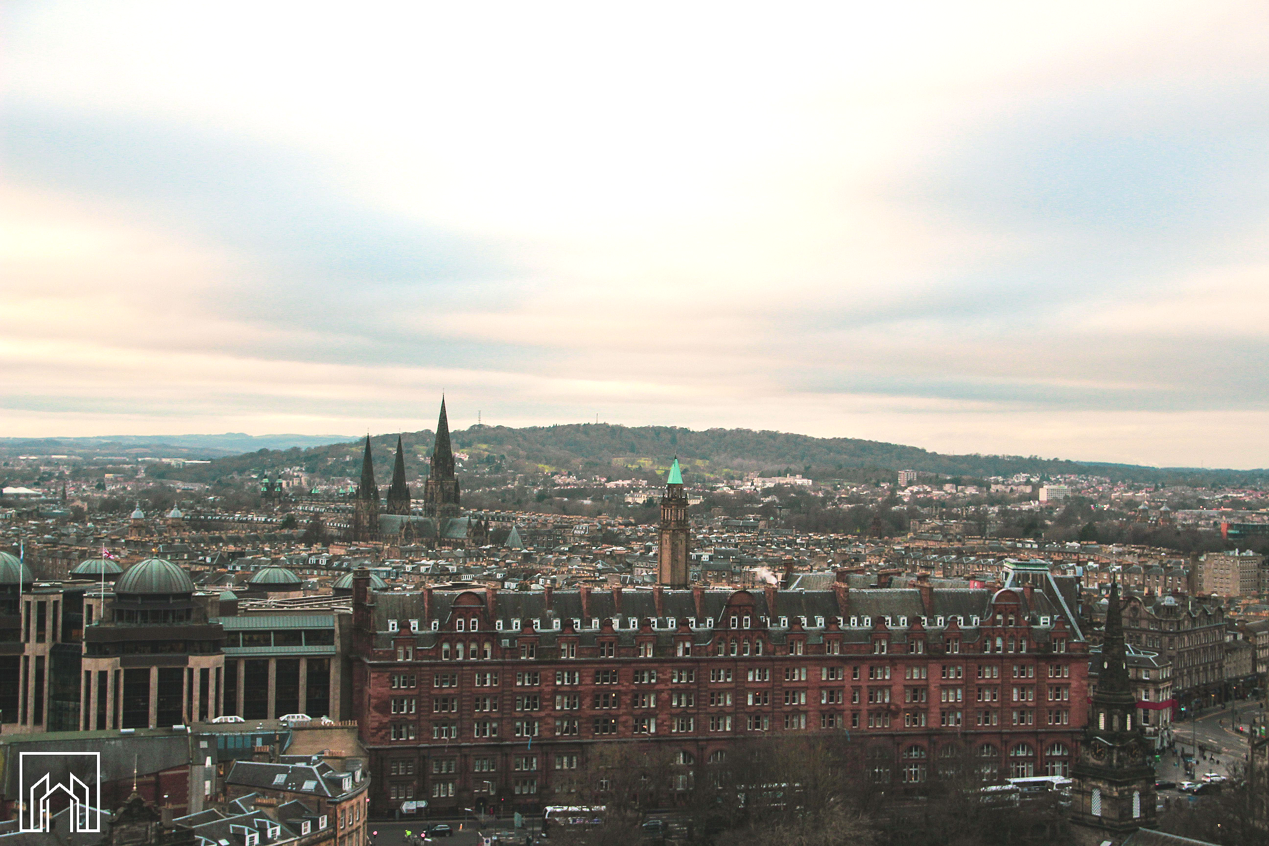 A cityscape of a historic European city, with gothic cathedral spires and red brick buildings in the foreground, and a hilly landscape in the background under a cloudy sky.