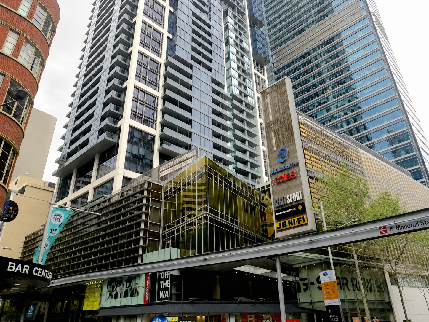 A city street view with tall modern glass and steel buildings, and signs for Melbourne, Coles, Rebel Sport, JB Hi-Fi, and a Monorail station entrance.