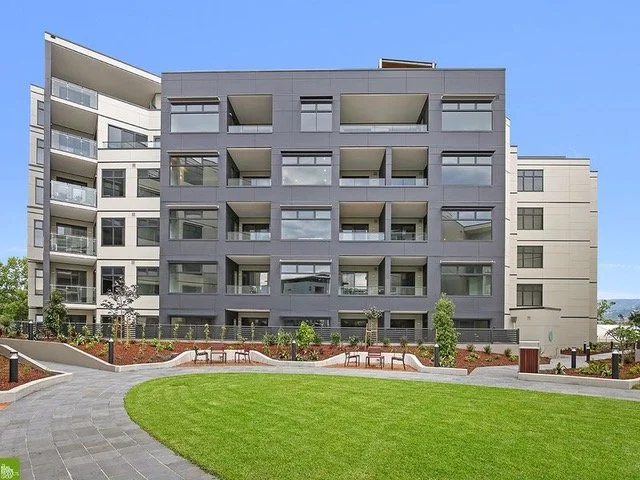 Modern multi-story apartment building with balconies, situated behind a landscaped lawn with benches in an urban setting.
