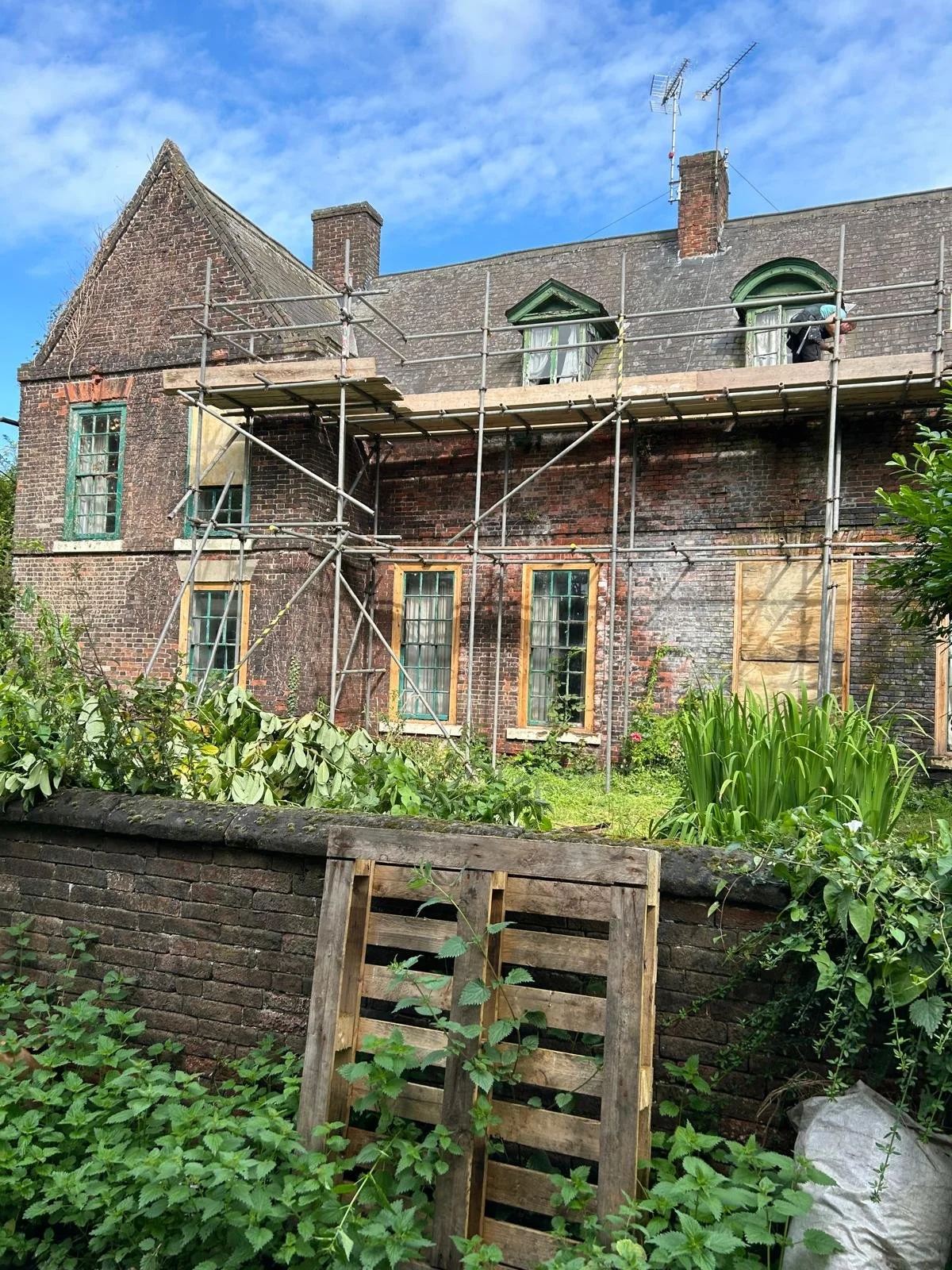 An old brick house under renovation with scaffolding around it. Windows are visible, some covered with plastic or tape. There is greenery in the garden below, and a wooden pallet leaning against a brick wall in the foreground.