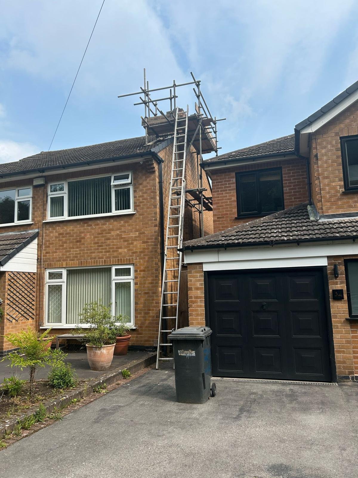 A brick two-story house with a black garage door, a ladder leaning against the house, and scaffolding on the roof, indicating roof repair or maintenance work.