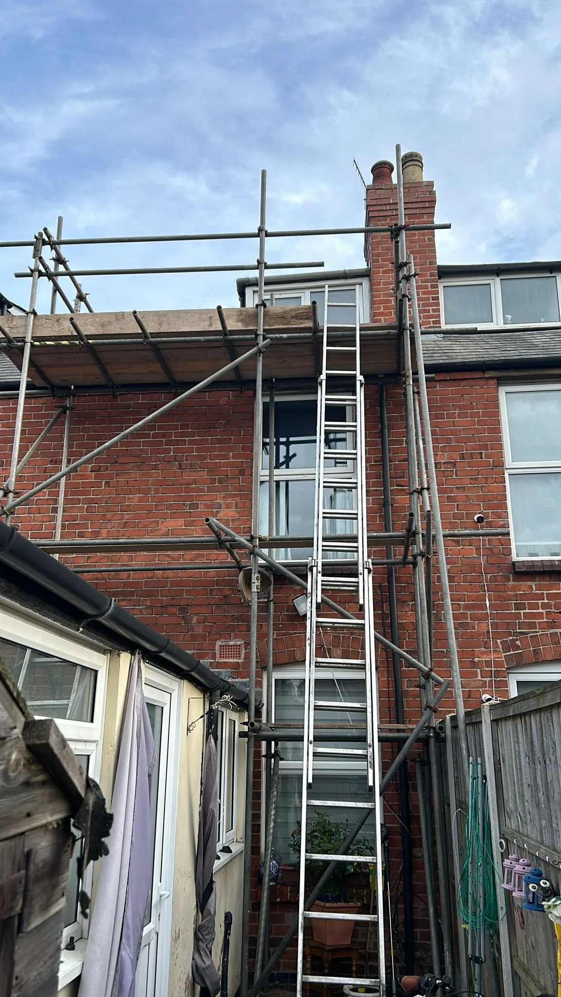 Residential house with brick exterior, scaffoldings, ladder, and chimney under a partly cloudy sky.