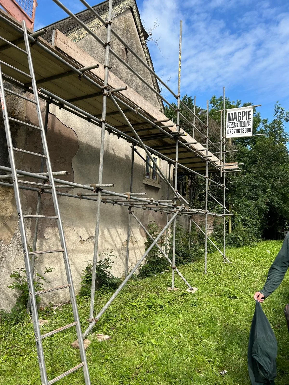 A building with scaffolding around it, showing signs of renovation or repair, with a sign that reads 'MAGPIE SCAFFOLDING' and a phone number. The building's exterior wall appears worn with visible cracks, and there is greenery and trees in the backgr