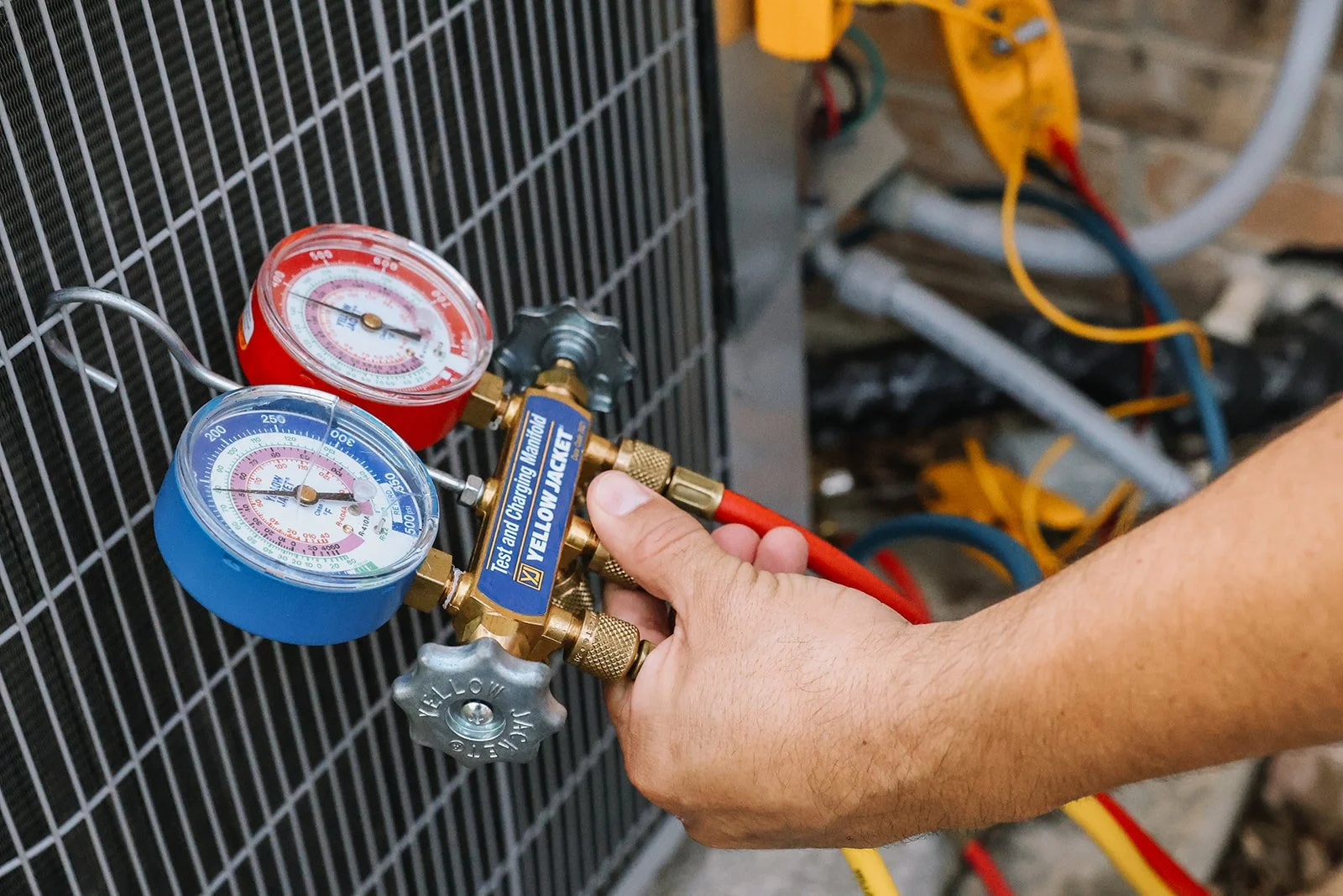 A technician's hand holding a manifold gauge set attached to an air conditioning unit, with pressure gauges, hoses, and valves visible.