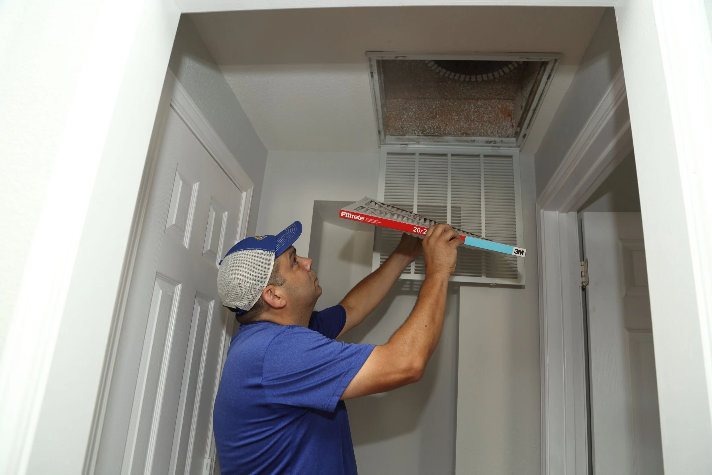 A man wearing a blue shirt and a cap is installing a new air vent cover in the ceiling of a small room.
