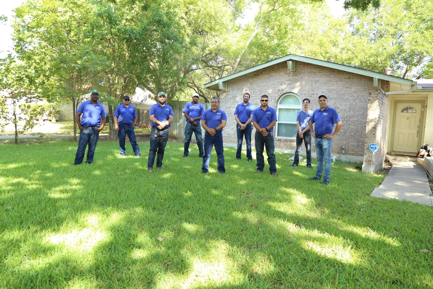 A group of men in blue shirts standing on a green lawn in front of a house with brick exterior and large trees overhead