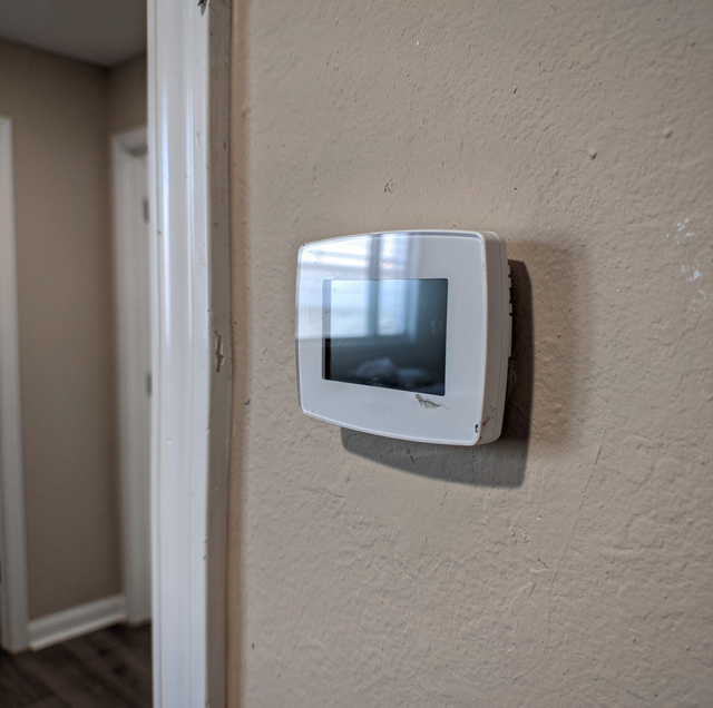 A white thermostat with digital display mounted on a textured beige wall next to a white door frame.