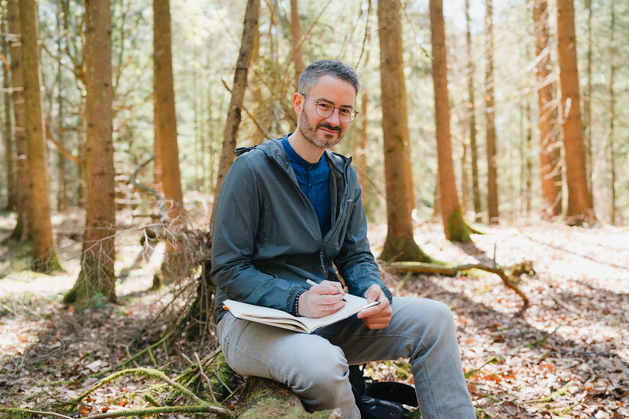 Mann sitzt im Wald auf einem Baumstamm, schreibt in ein Notizbuch, trägt eine graue Jacke und eine blaue Shirt, umgeben von Bäumen und herbstlichen Blättern.