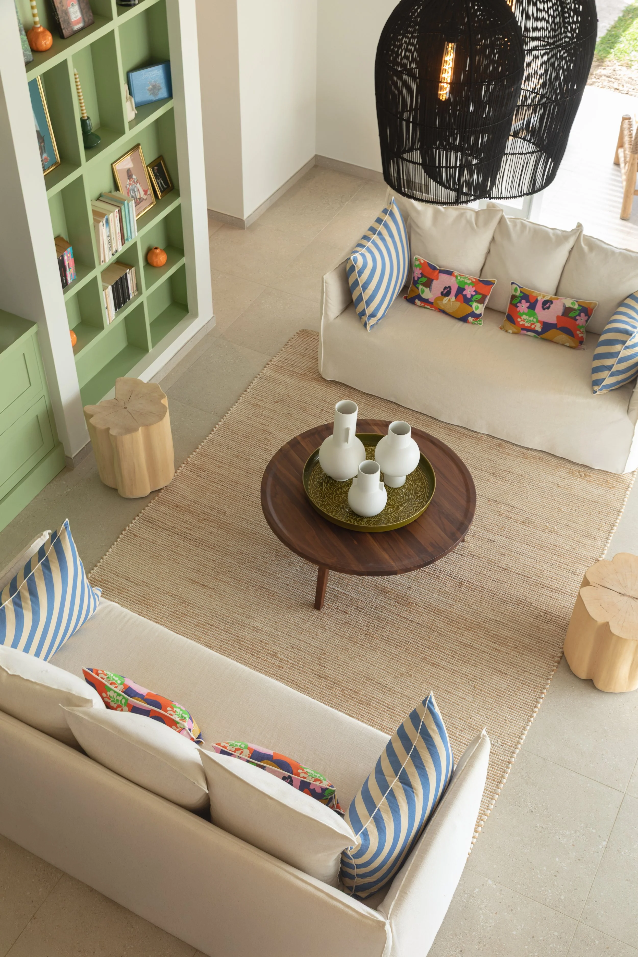 Vue aérienne d'un salon avec un canapé beige et des coussins colorés, une table ronde avec des vases blancs, une étagère verte avec des décorations et livres, un luminaire noir suspendu, et un tapis en jute sur un sol en carreaux