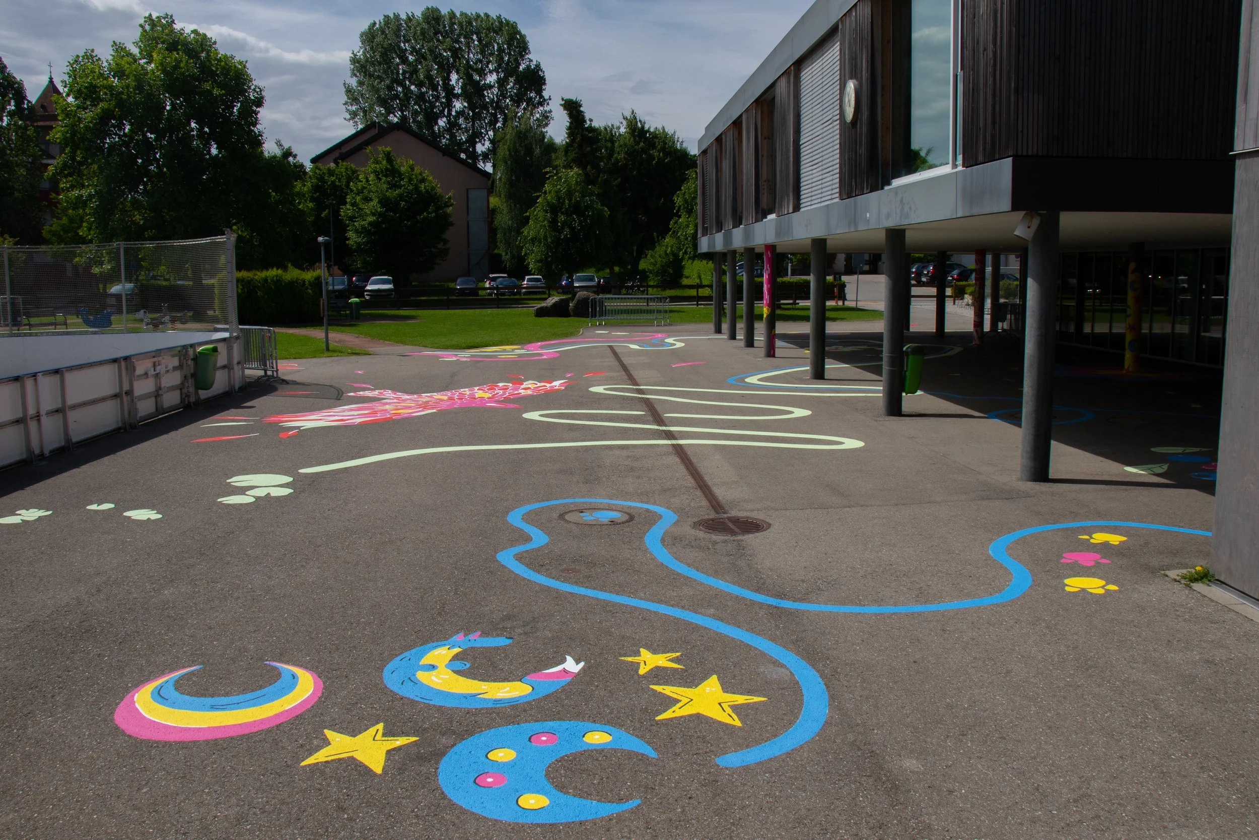 Cour d'école colorée avec motifs de dessin de comètes et étoiles, entourée d'arbres et de bâtiments, sous un ciel partiellement nuageux.