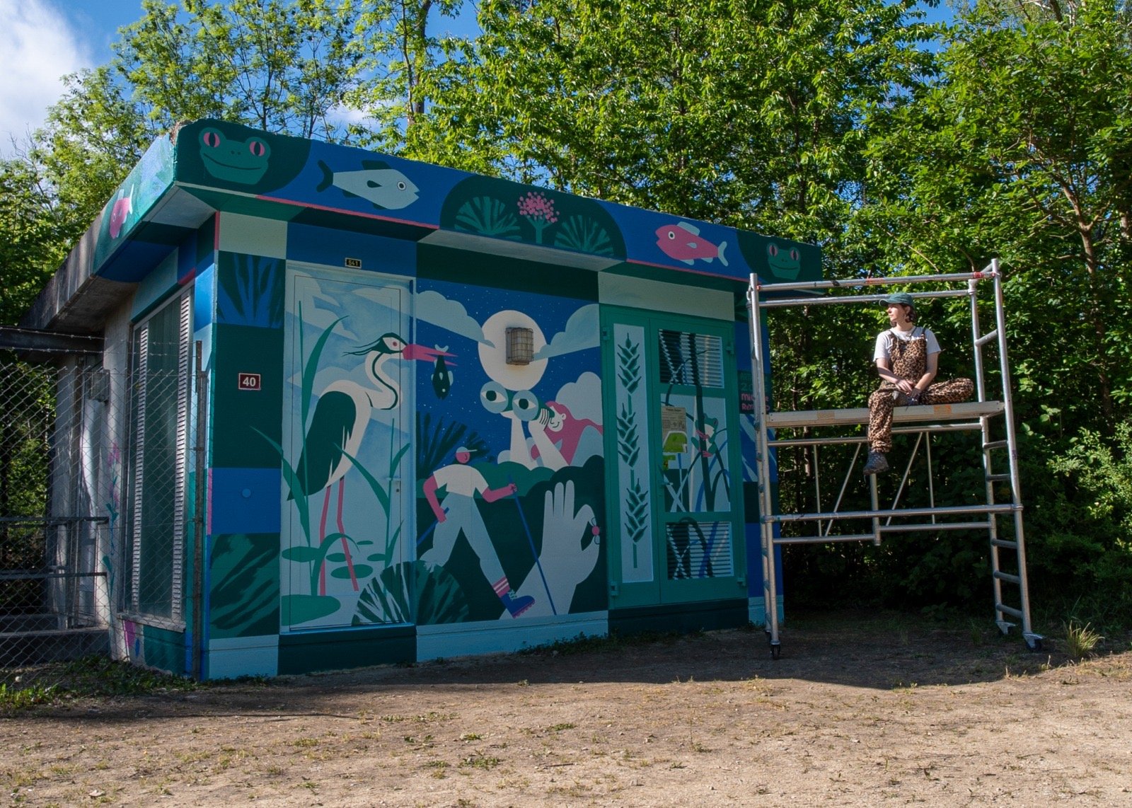 Murale colorée représentant des animaux et des personnes dans un style artistique, avec une personne assise sur une échelle à côté de l'œuvre, dans un environnement extérieur avec des arbres en arrière-plan.