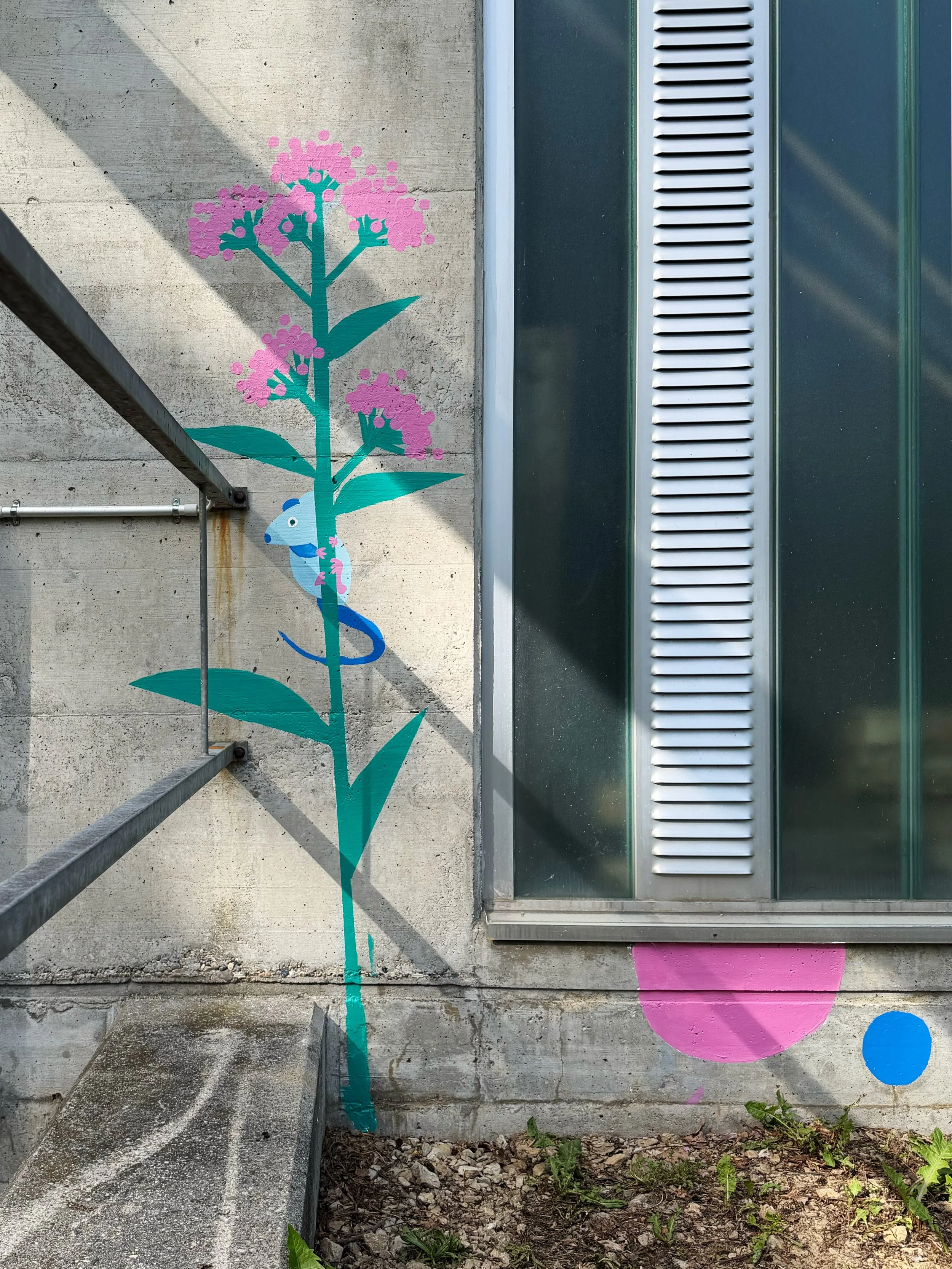 Une fresque murale représentant une plante avec des fleurs roses, une souris bleue est accrochée à la tige, le tout peint sur un mur en béton à côté d'une fenêtre ou ventilateur en métal.