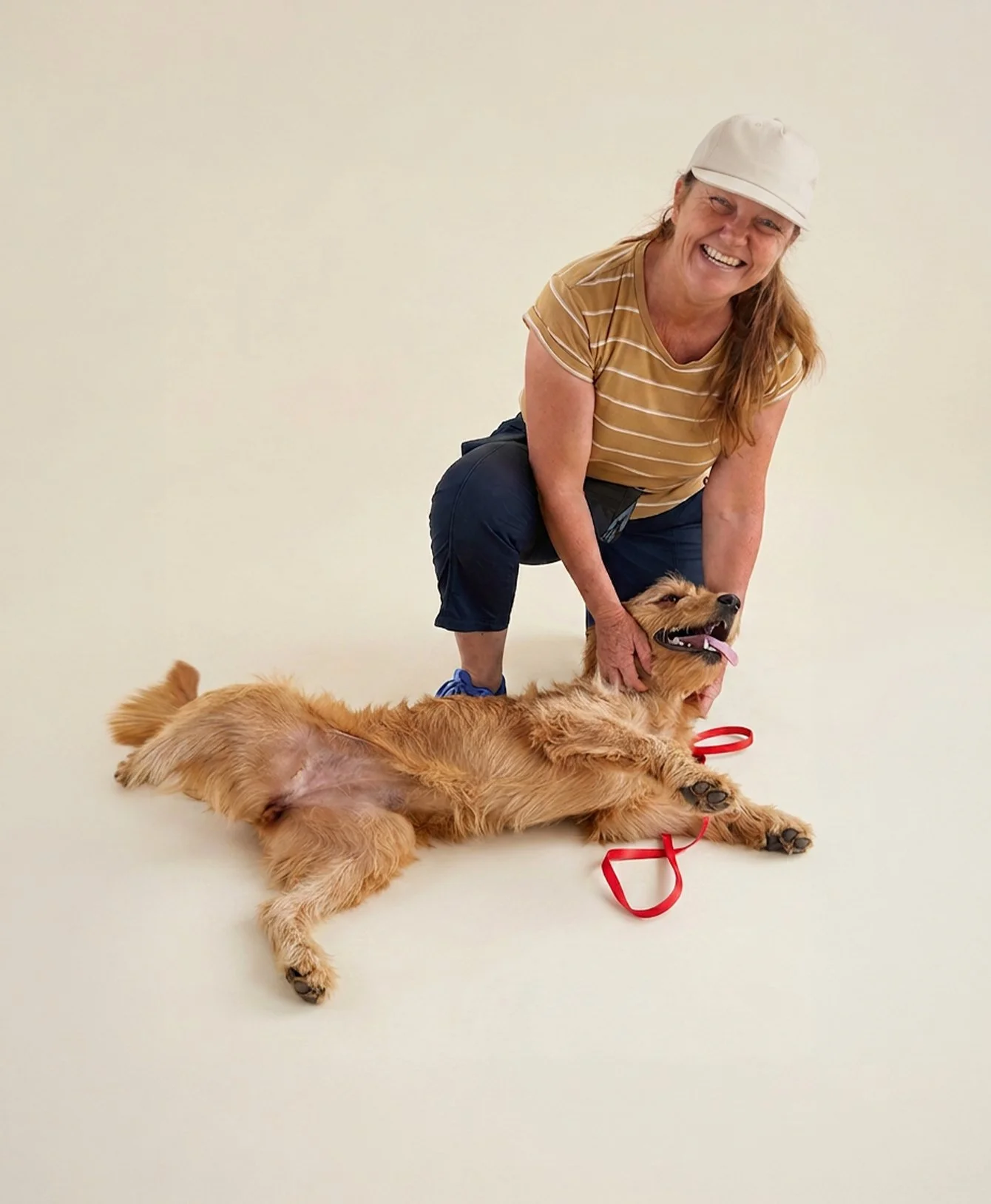 A smiling woman with long red hair, wearing a beige cap, striped yellow shirt, and navy pants, kneeling next to a large golden retriever lying on its back with a red leash, against a plain off-white background.