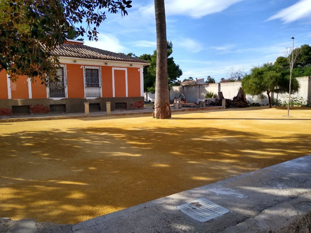 A small orange house with white trim and barred windows. There is a large tree in the yard with shadows on the ground, and some additional trees and bushes along the walls. The yard appears to be freshly covered with yellow material, possibly gravel or mulch. The sky is blue with some clouds.
