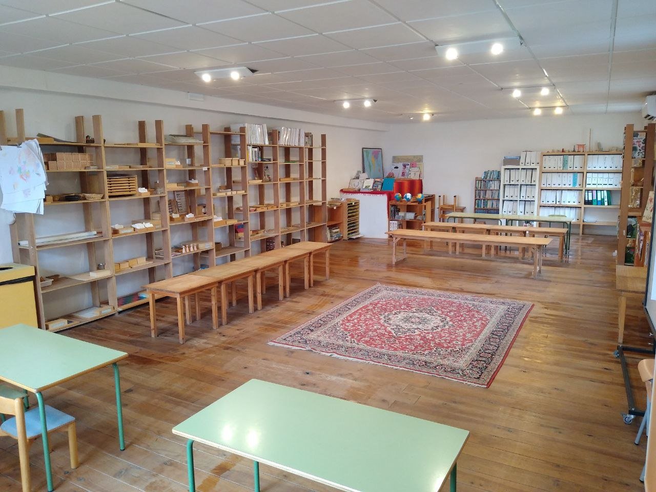 Empty classroom with wooden shelves, tables, and chairs, a colorful rug, and educational materials on the shelves.