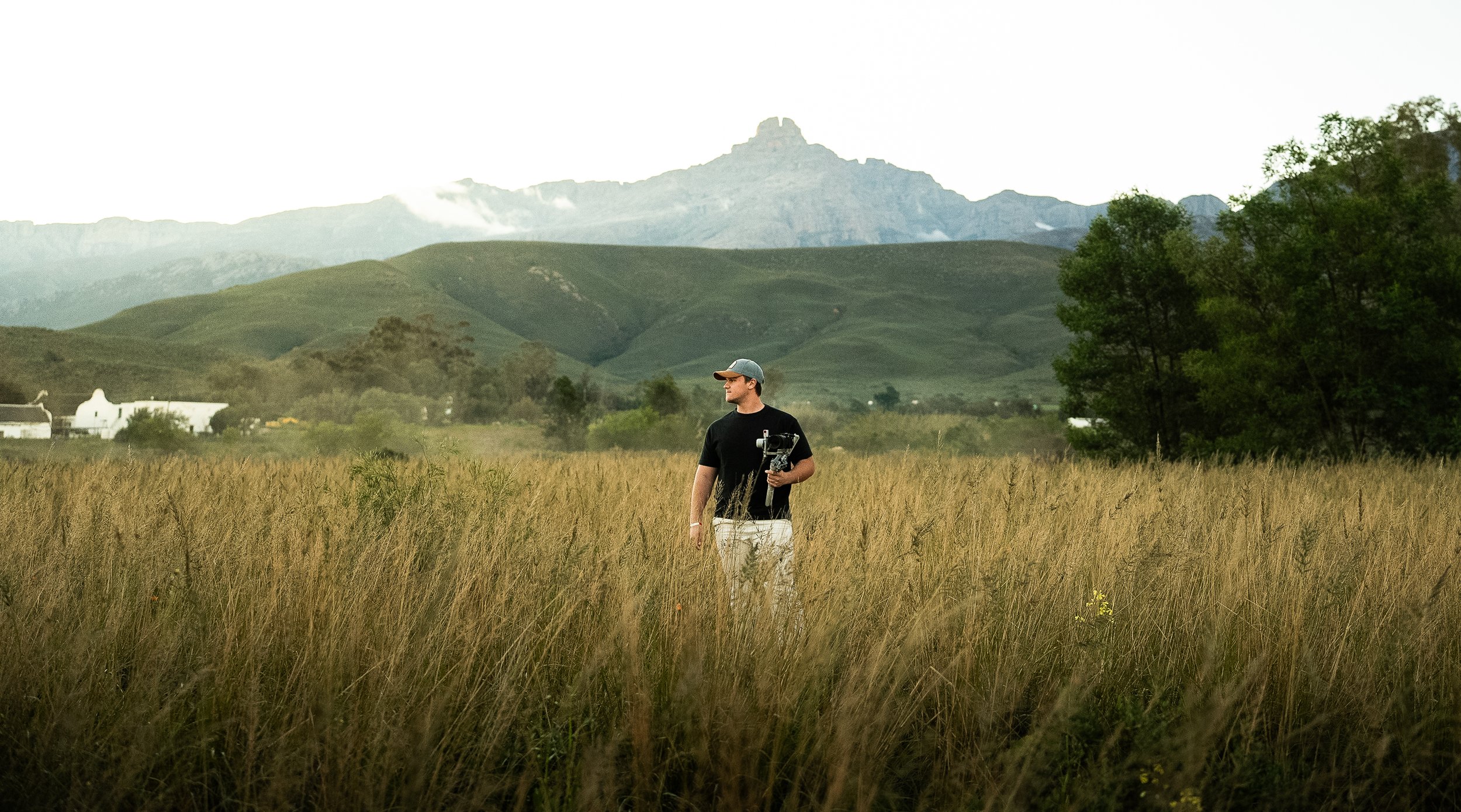 A man standing in a grassy field with mountains in the background, holding a camera mounted on a stabilizer, wearing a baseball cap and glasses.