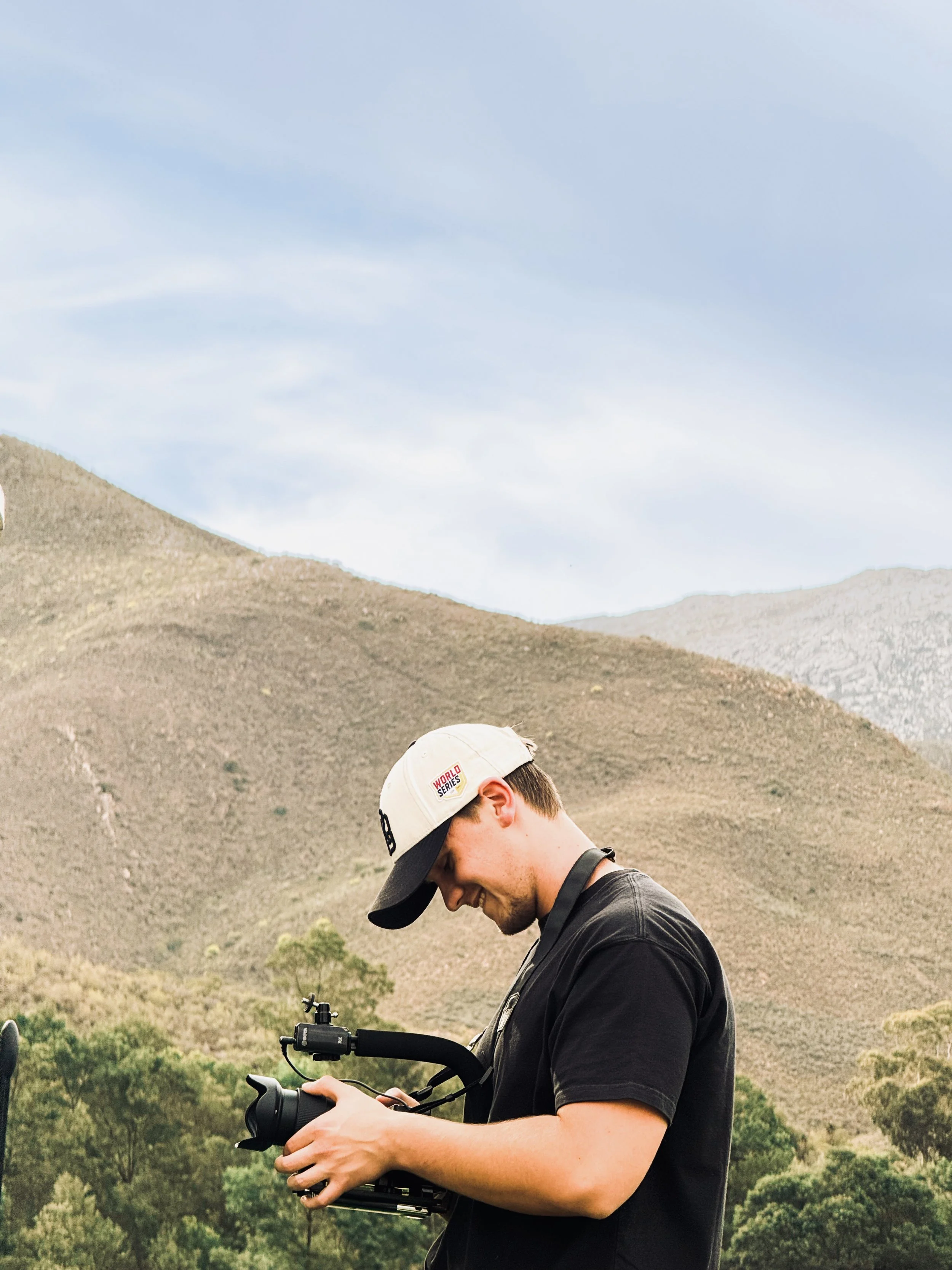 A young man outdoors holding a drone controller, wearing a beige baseball cap and a black t-shirt, with a mountainous landscape and trees in the background.