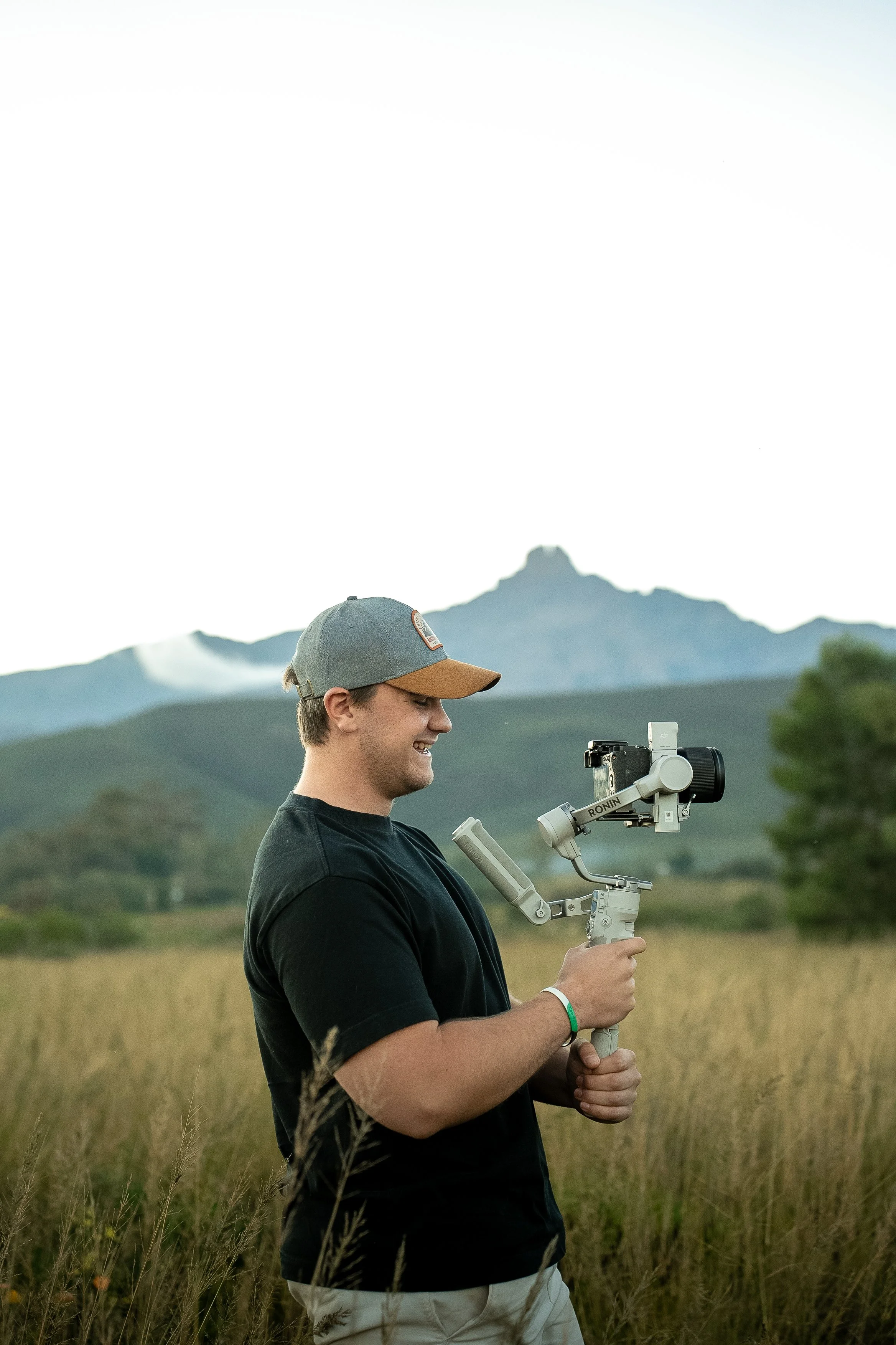 A young man in a black t-shirt and beige pants holding a camera stabilizer in a field with tall grass. He is wearing a gray cap and is smiling as he looks at his camera. Mountains and trees are visible in the background.