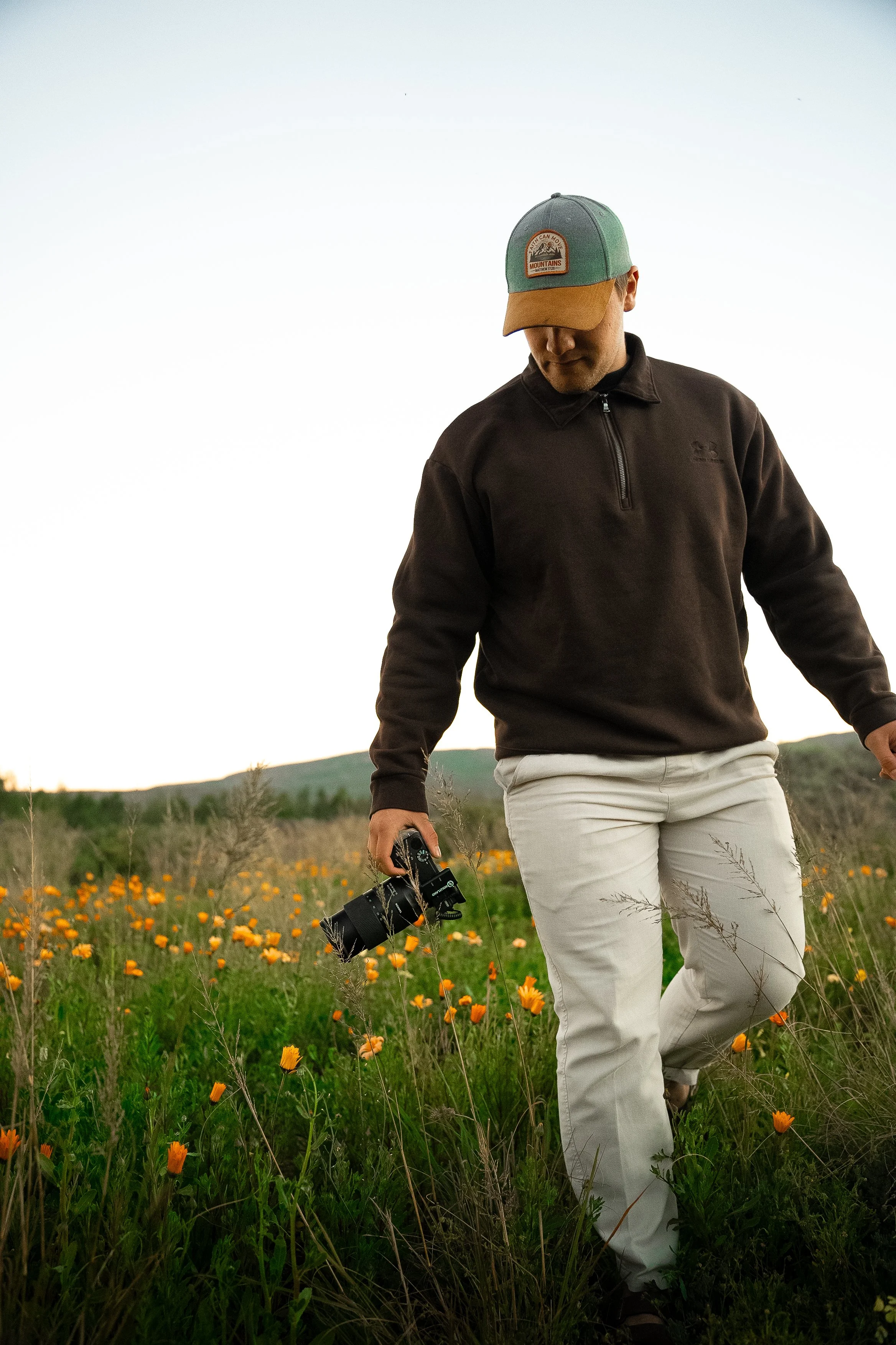 A person is walking through a field of orange flowers, wearing a black zip-up jacket, beige pants, and a blue and brown cap, holding a camera in their right hand.