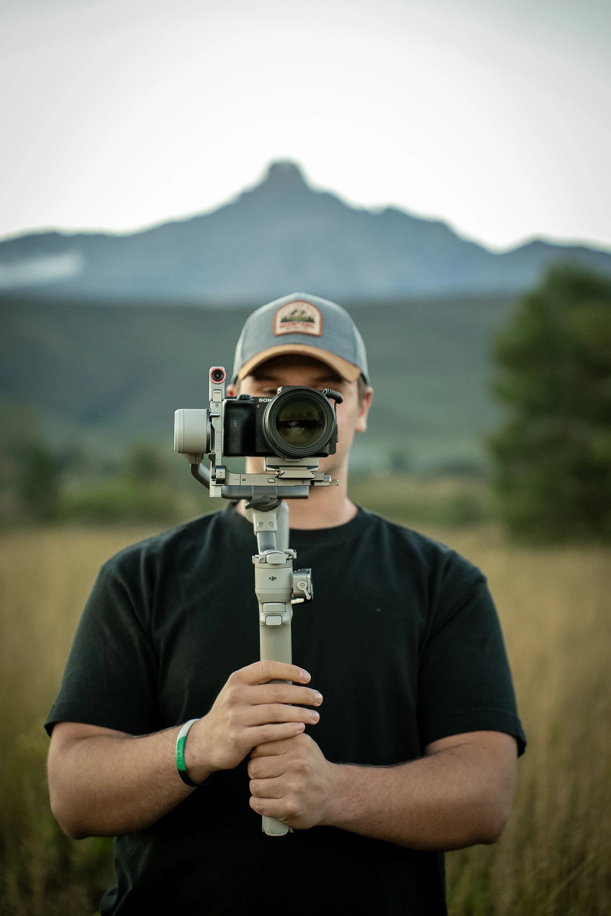 A person in a black shirt holds a camera on a tripod, facing forward, with a mountain in the blurred background.