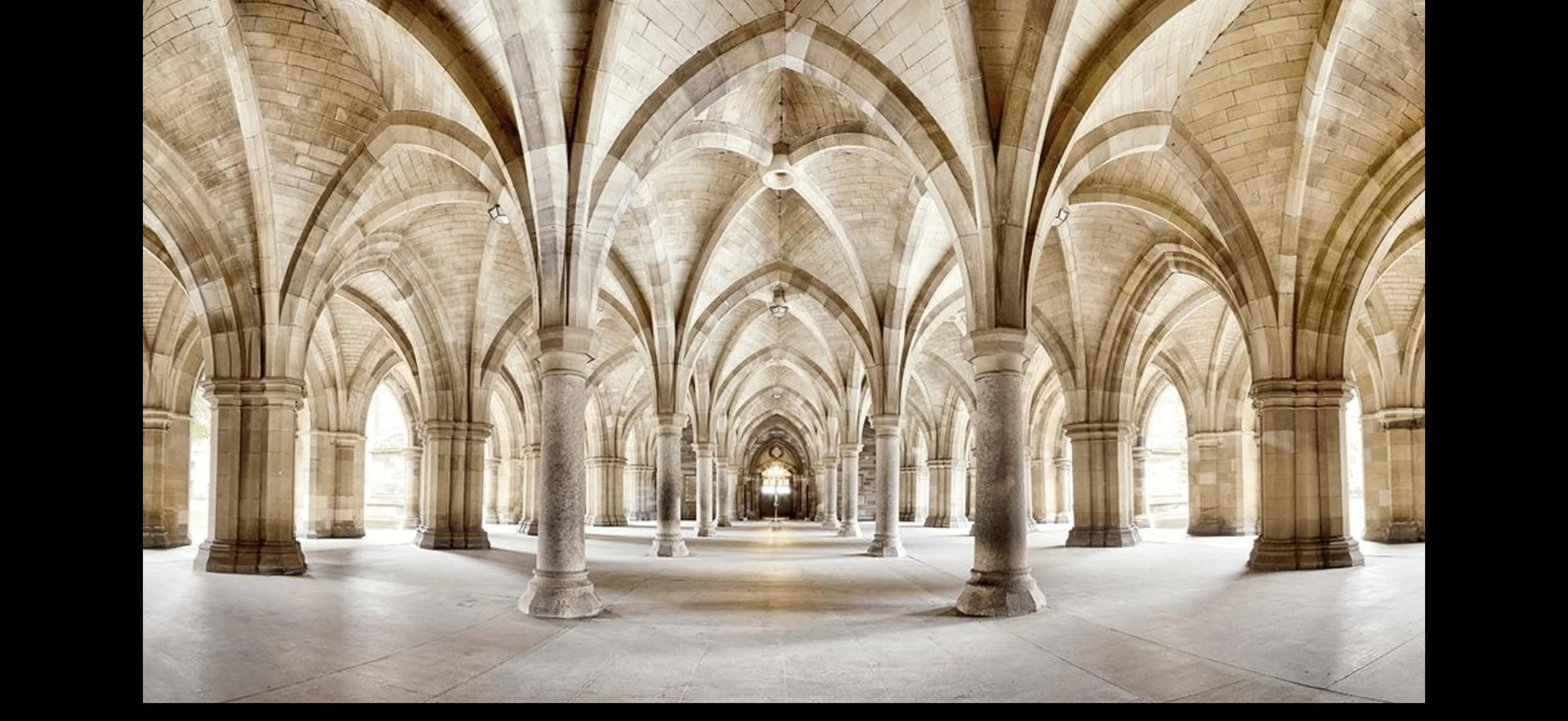 A grand stone hall with Gothic-style pointed arches supported by columns, featuring high vaulted ceilings and natural light flooding in through large windows.