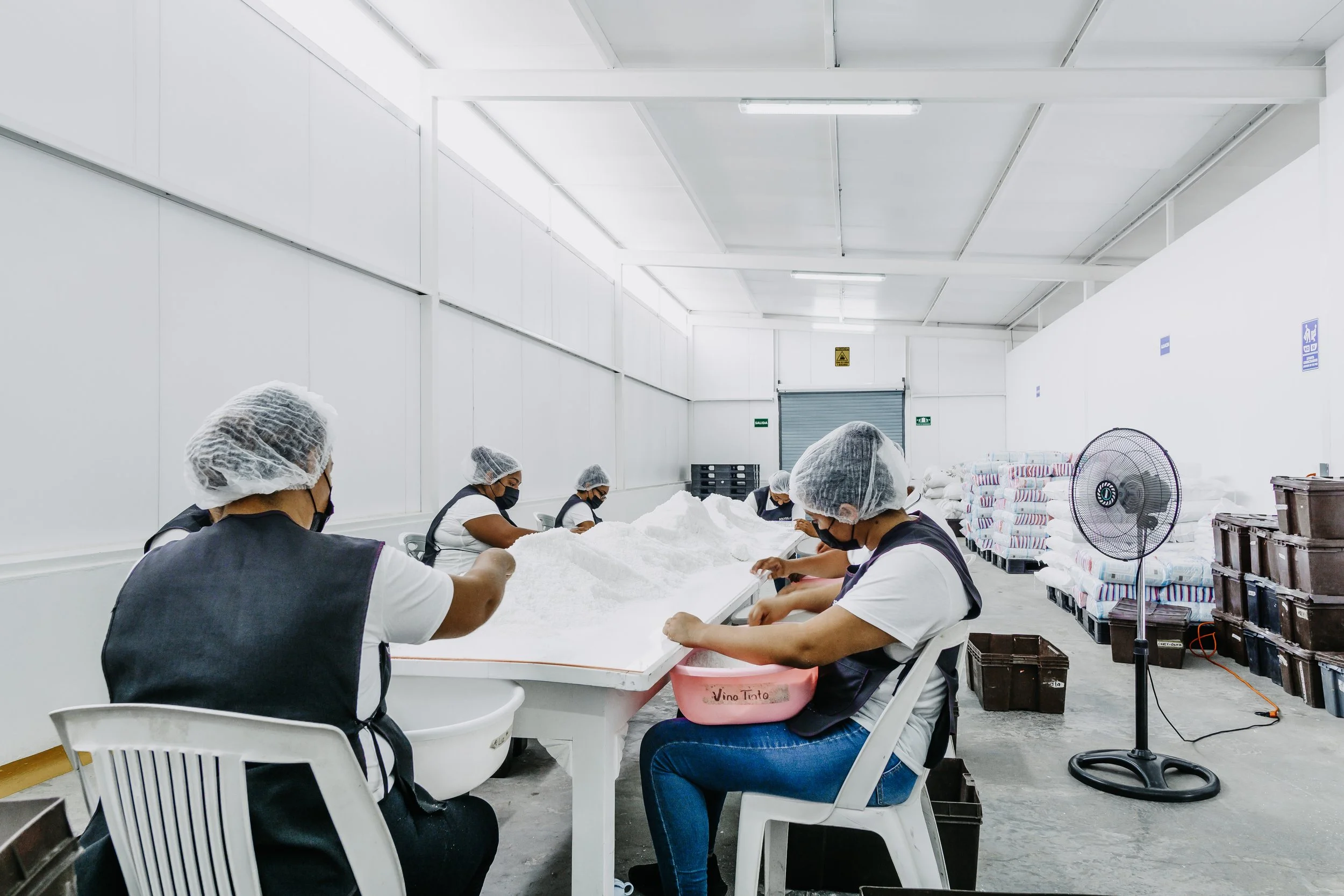 Trabajadores en una fábrica empacan productos en una mesa blanca, con gabinetes y señales en la pared, con un ventilador de pie y bolsas apiladas en el fondo.
