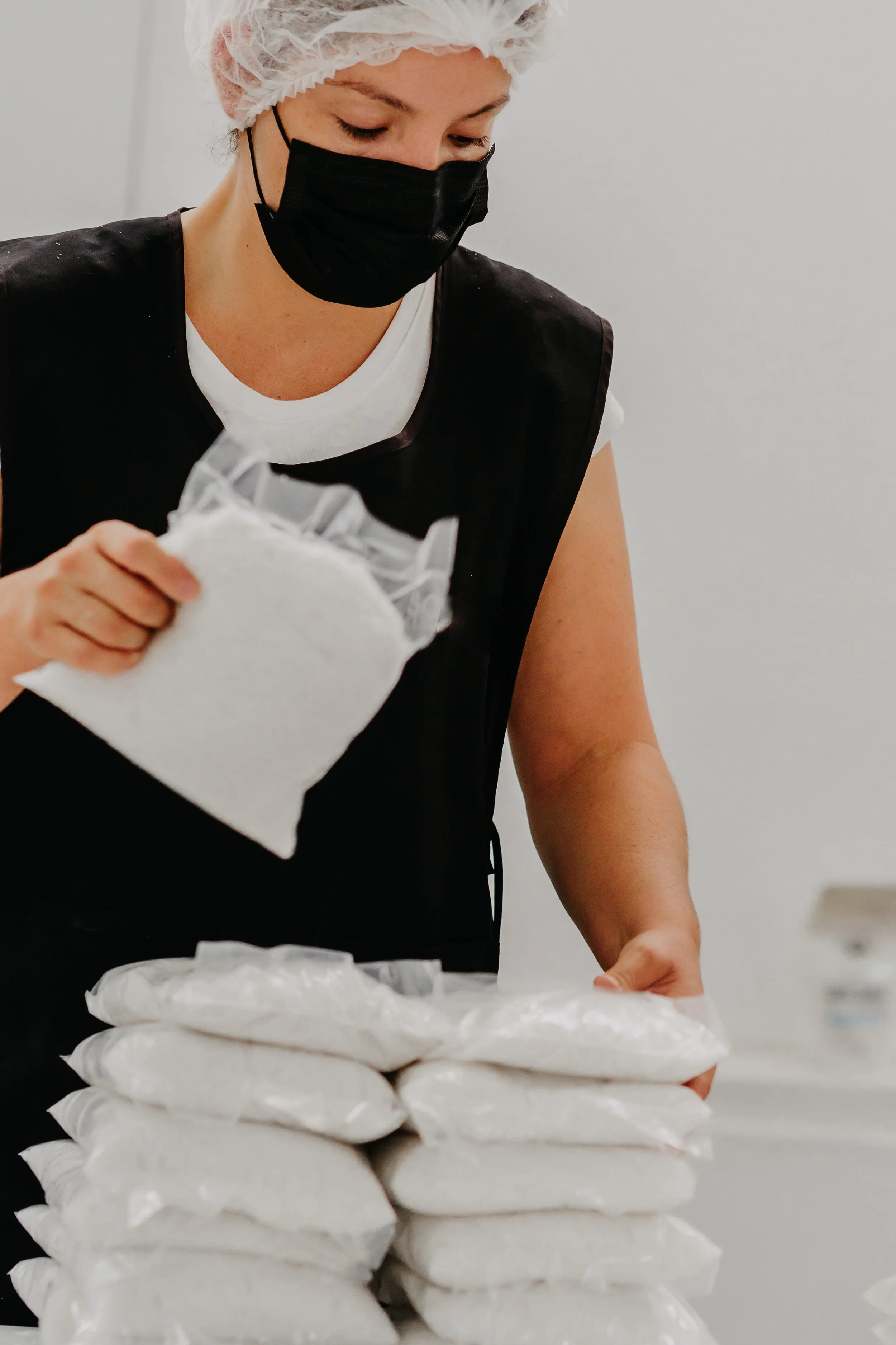 Mujer con gorro y mascarilla negra, trabajando con paquetes de algodón en una superficie blanca.