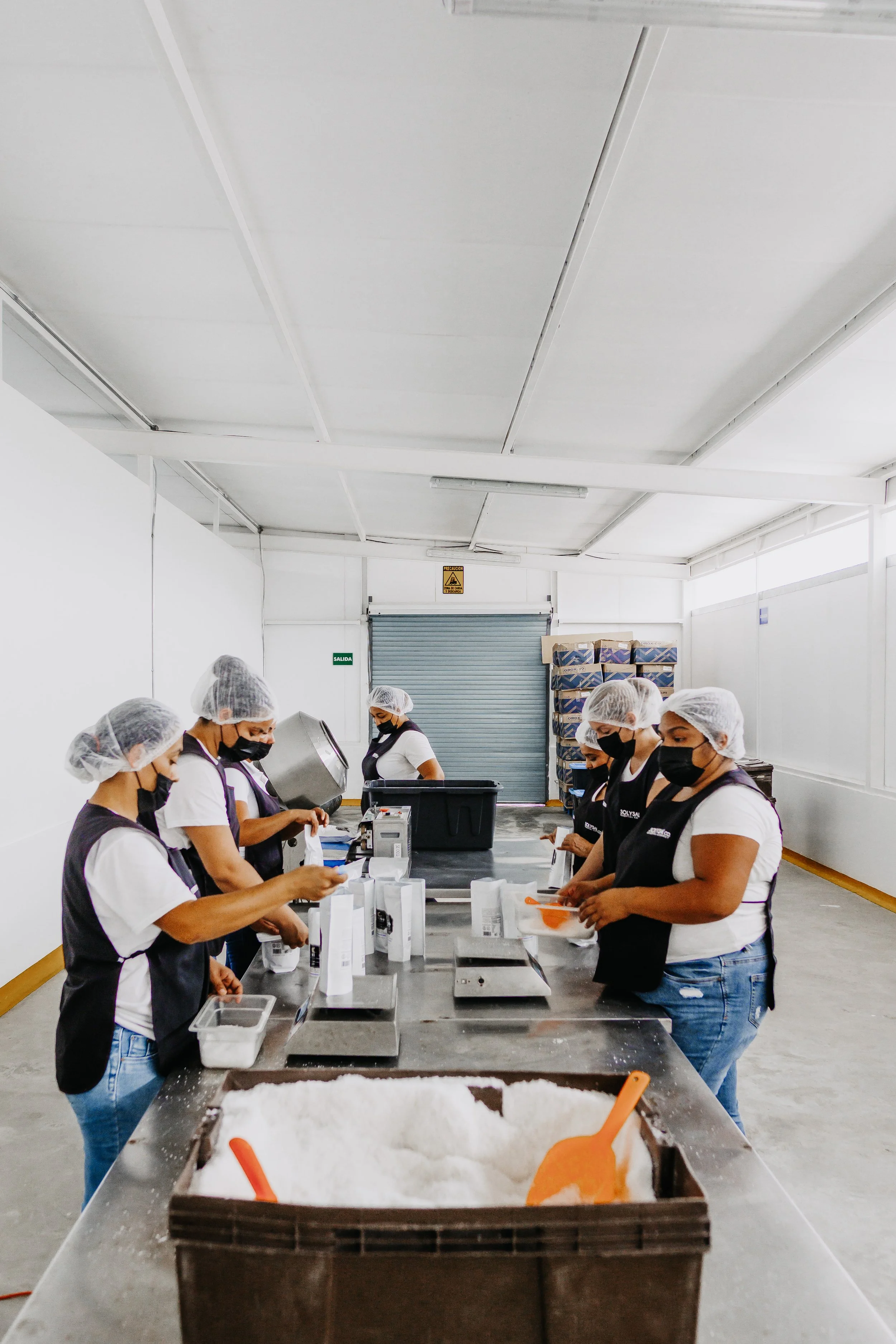 Personas trabajando en una línea de producción en un ambiente industrial, con equipo de protección y mascarillas, empacando productos en bolsas de papel. En el fondo, se observa una cortina de metal y unas estanterías con cajas y un cartel de precauc