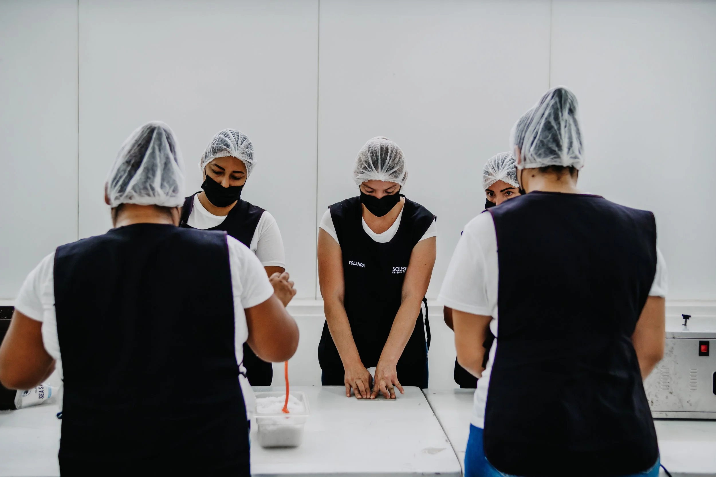 Grupo de personas en una cocina, con batas, gorros de plástico y mascarillas, hacen panadería o repostería, concentrados en su trabajo.