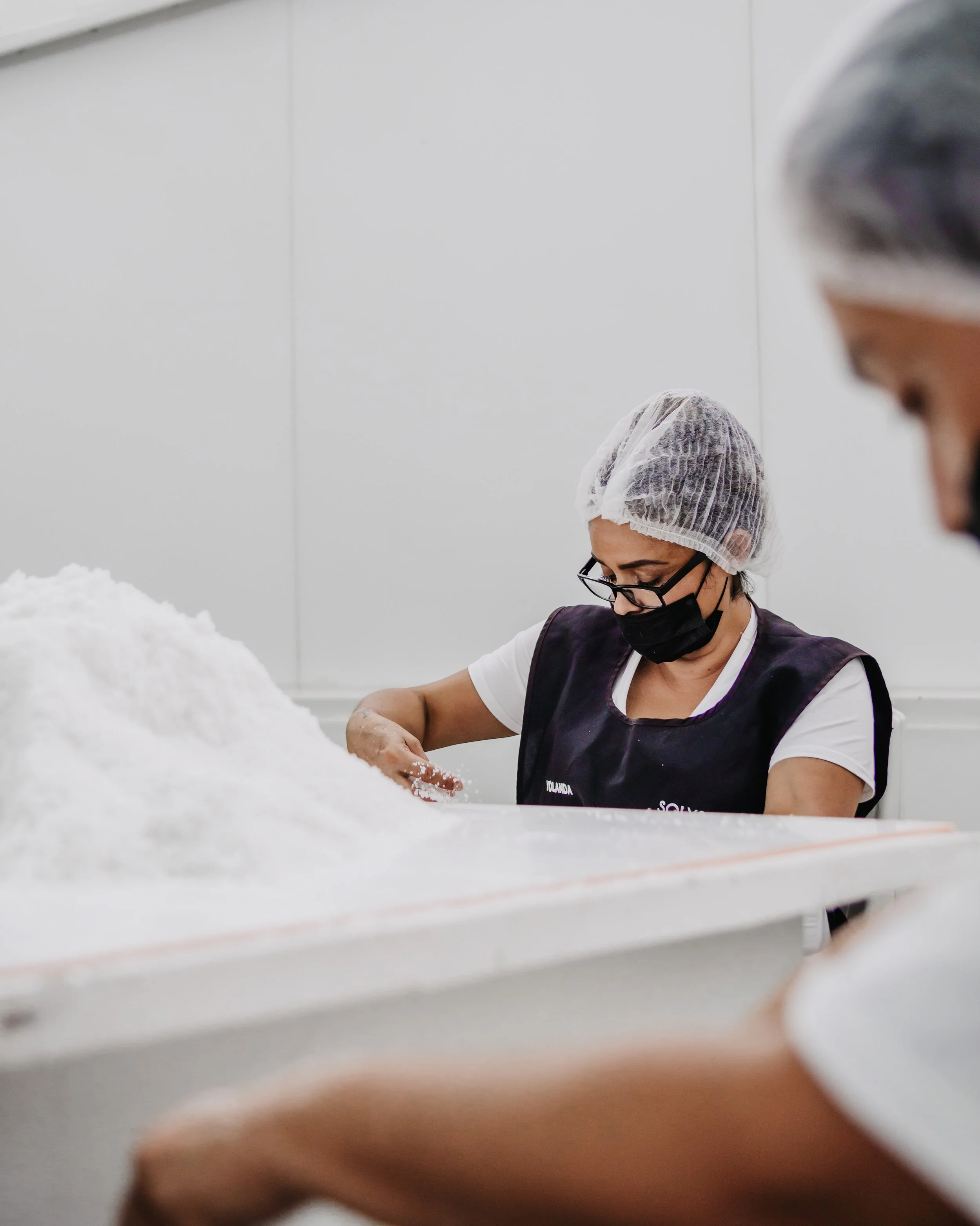 Una mujer con gafas y máscara negra, usando gorro de red y uniforme, trabaja con algodón en un ambiente de producción.