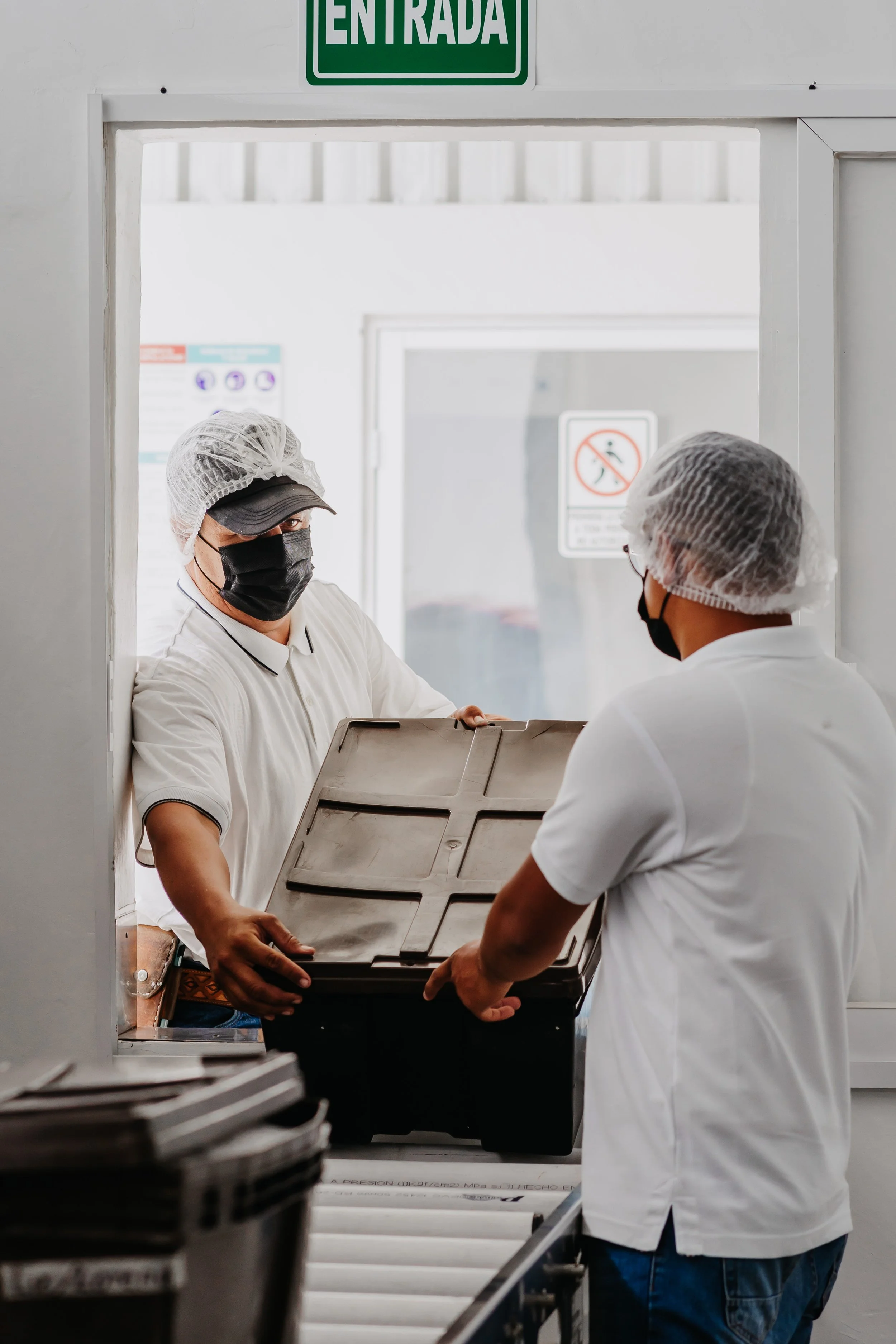 Dos empleados usando gorros, mascarillas y pantalones cortos, trabajan en una estación de comida o café, entregando un contenedor con alimentos a través de una ventana de servicio.