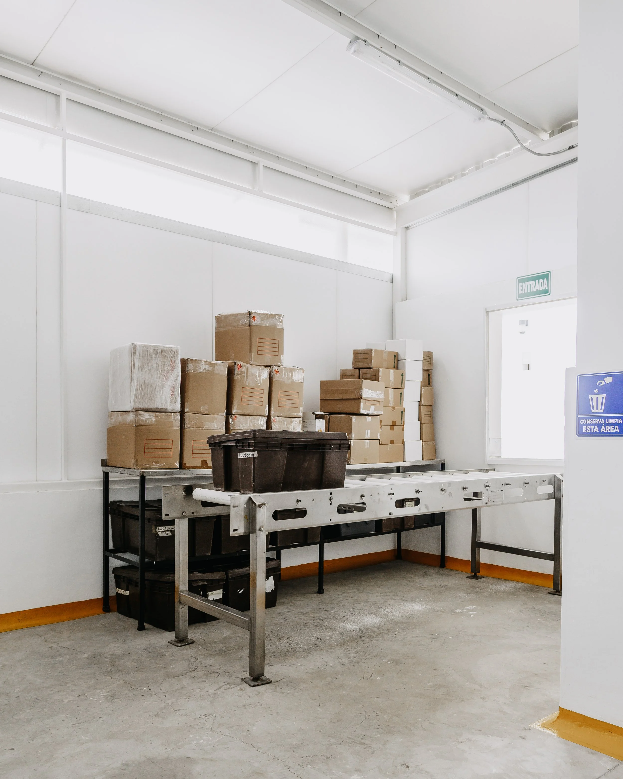 Interior de una bodega con cajas apiladas y mesa de trabajo, señal de entrada y símbolo de basura en la pared.