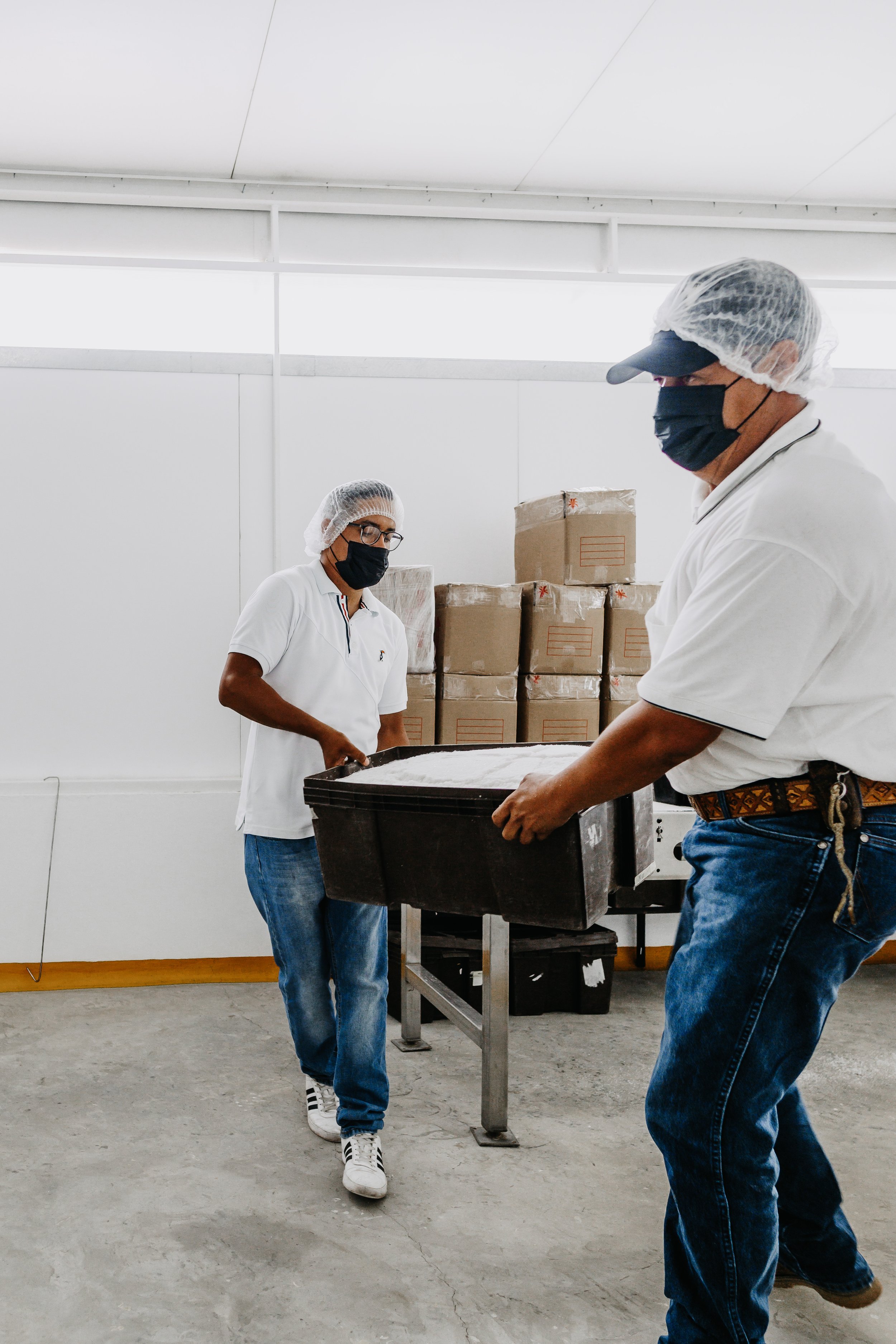 Tres personas en una planta procesadora de alimentos, con uniformes, gorros y mascarillas, manejando una bandeja grande con ingredientes de harina o masa, en un ambiente industrial con cajas apiladas al fondo.