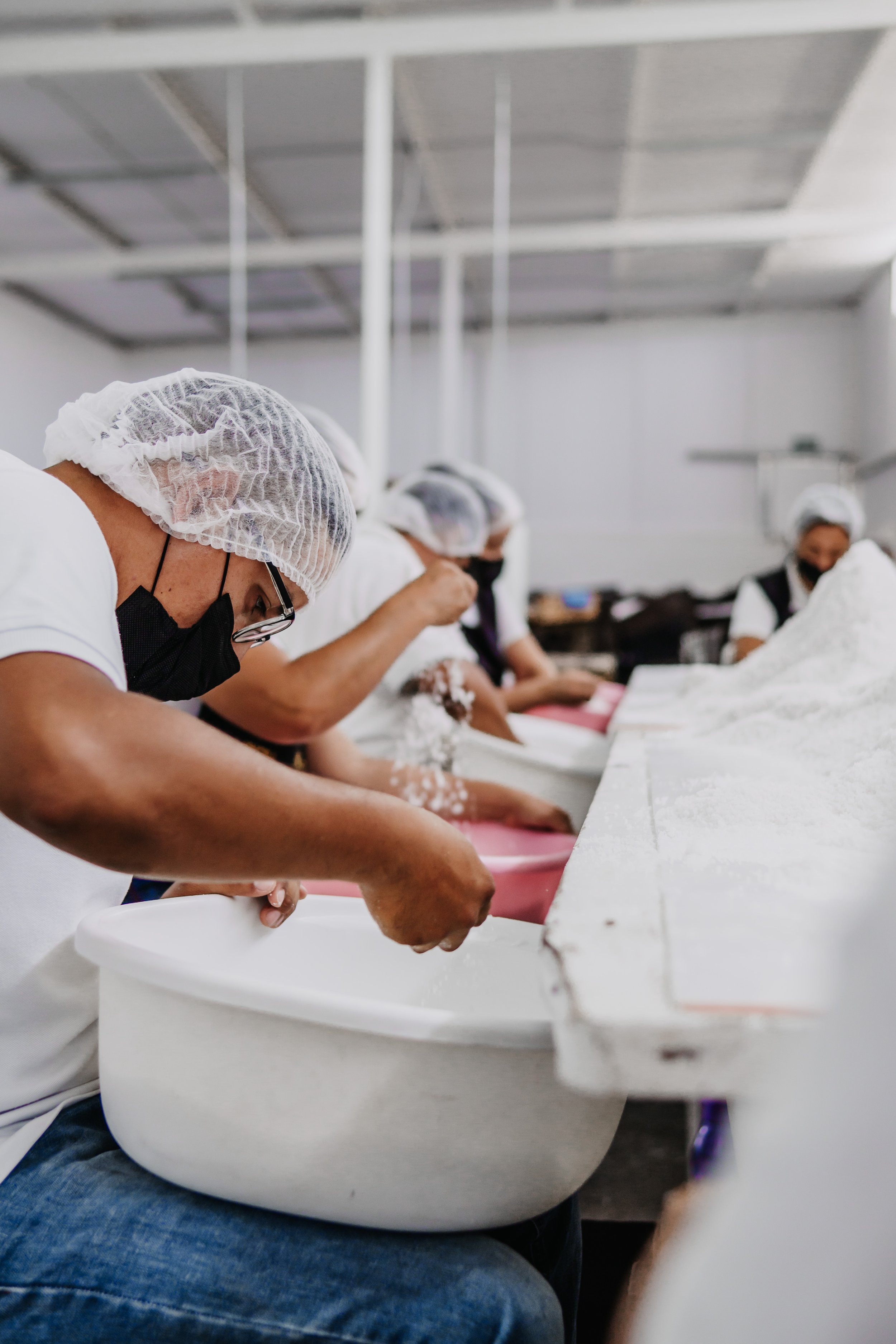 Personas trabajando en línea de producción, usando gorro y mascarilla, ensamblando productos.