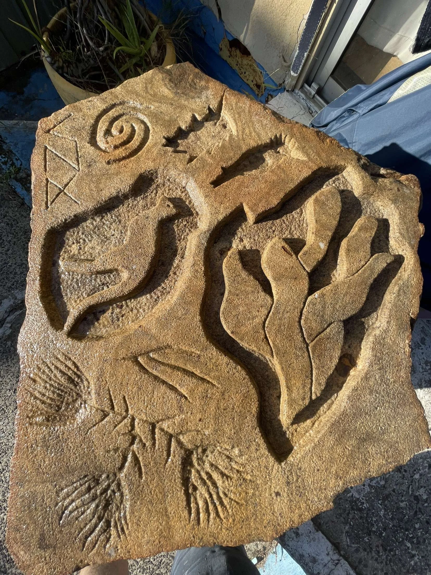 A carved stone with various spiral, linear, and geometric patterns, featuring a large spiral, jagged zigzag shapes, and an area resembling a leaf or petal, with a plant in a yellow pot and a sliding glass door in the background.