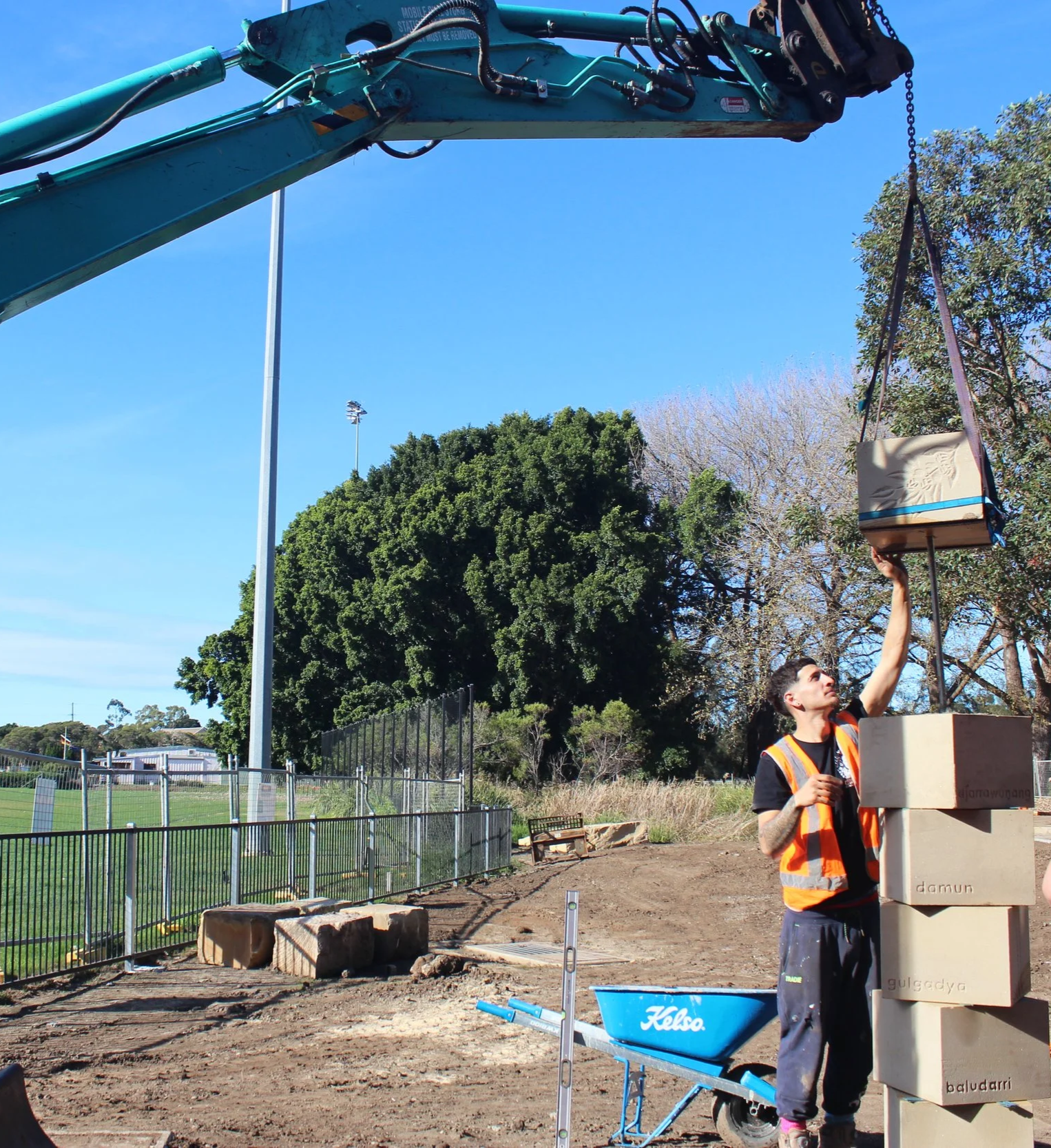 A person wearing a safety vest adjusting a large stacked box structure on a construction site outdoors. The background includes trees, a fence, a sports field with stadium lights, and a clear blue sky.