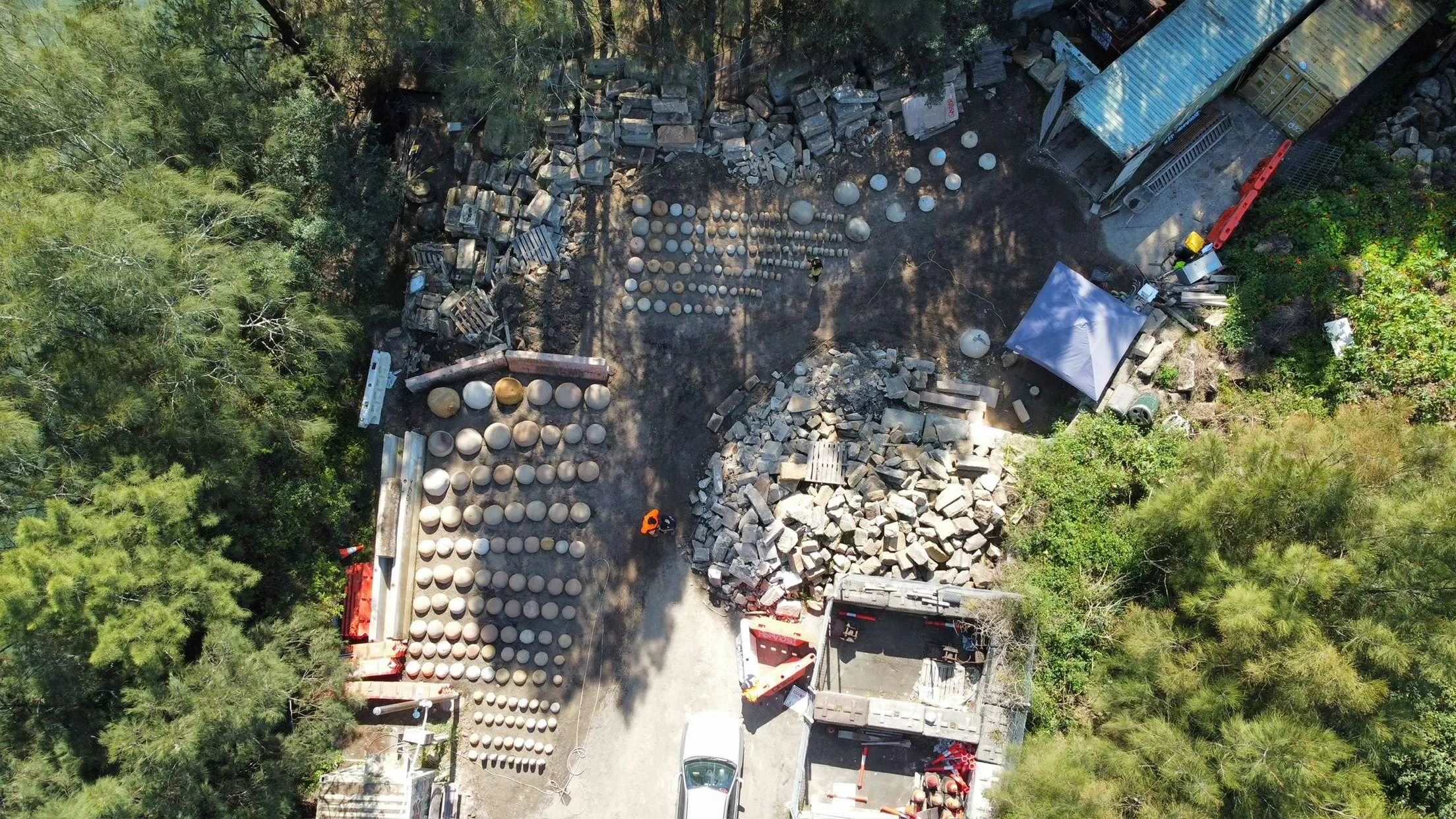 An aerial view of a construction or landscaping site with rows of large stones and spherical objects, surrounded by trees and green foliage, with workers and equipment present.