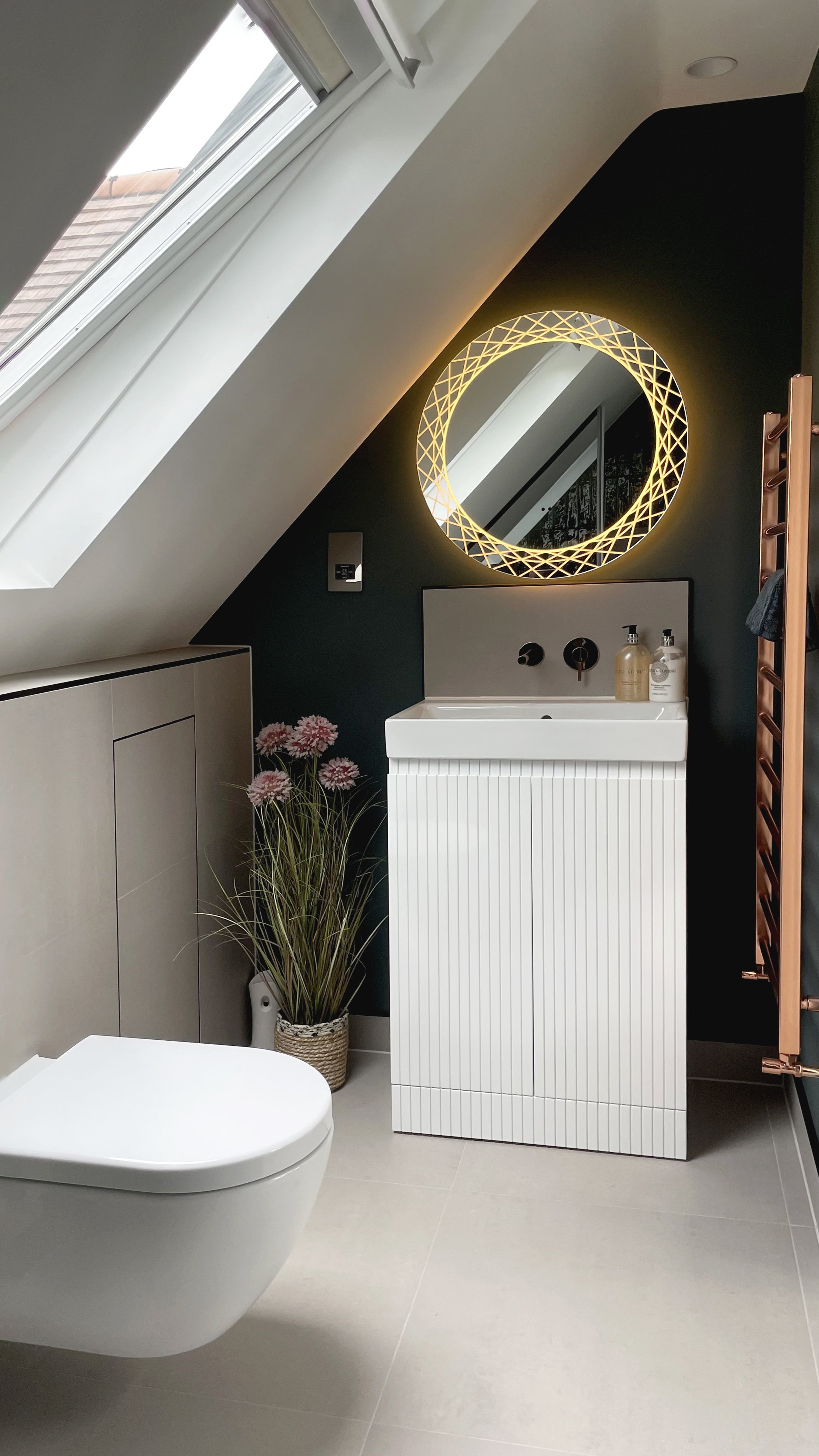 Modern bathroom with a sloped ceiling, skylight windows, a white toilet, a white sink with dark wall behind it, a round decorative mirror with lighting, a small cabinet, and a potted plant with pink flowers.