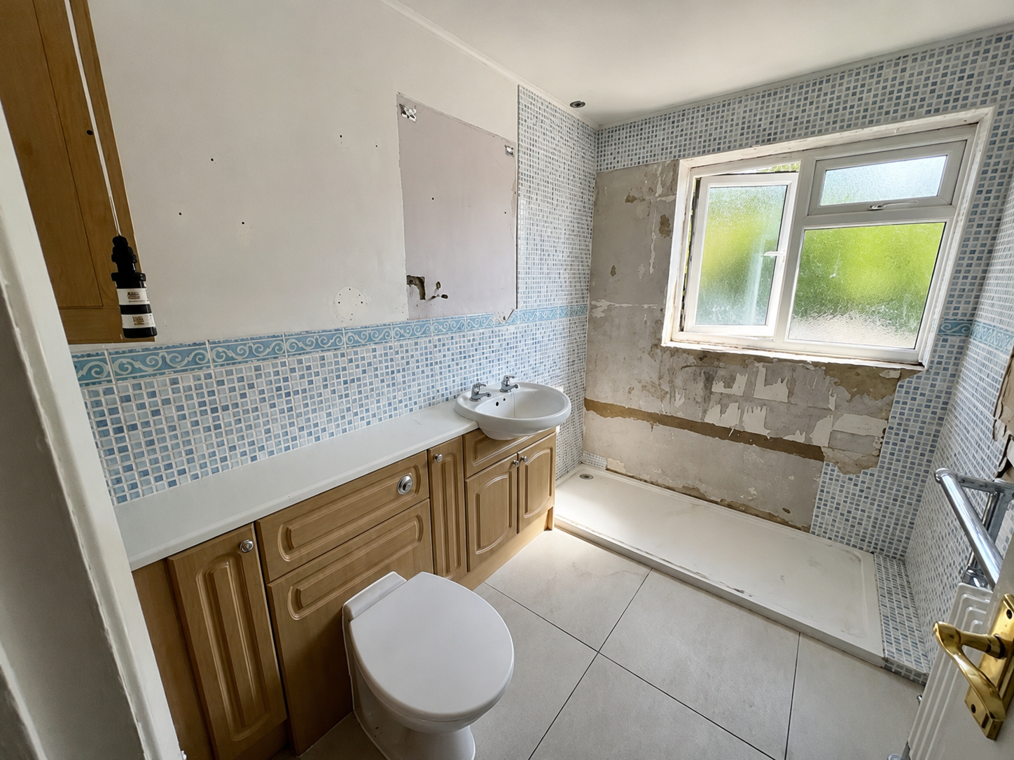 Bathroom under renovation with partially removed wall tiles, a window, a small white sink, and a toilet, with a shower area on the right side.