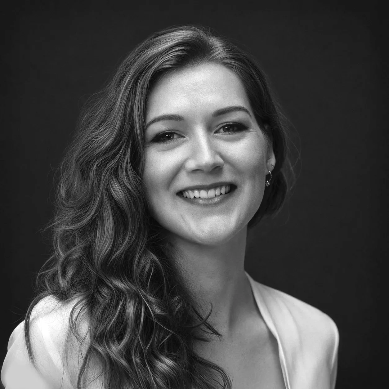 Black and white portrait of a smiling woman with wavy hair, wearing a light-colored top and earrings, against a dark background.