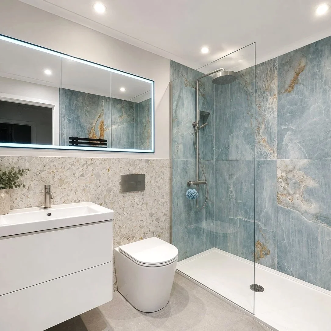 Modern bathroom with a white vanity, a potted plant, a wall-mounted toilet, a large illuminated mirror, a shower with blue marble tiles and a glass panel.