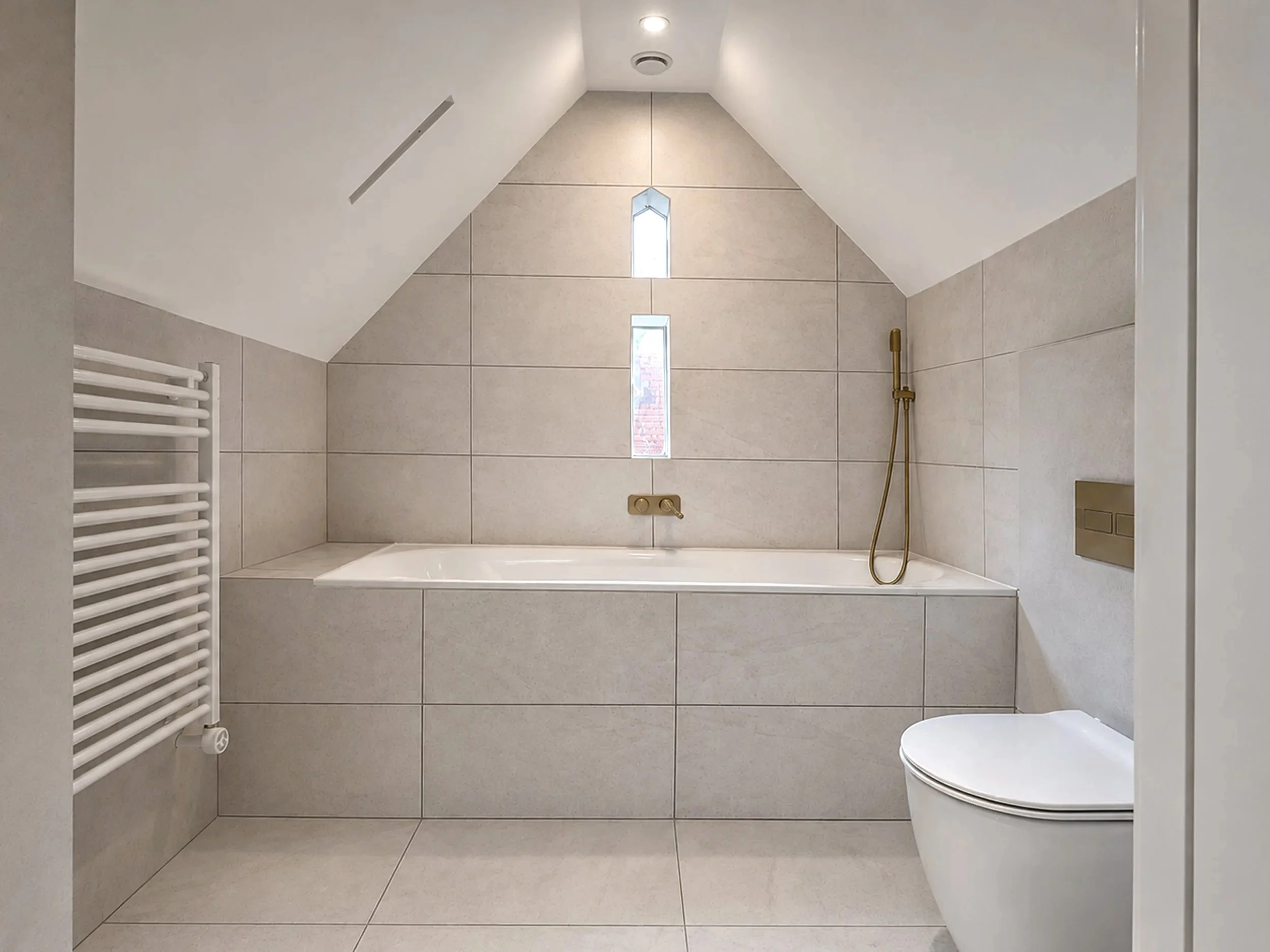 Modern minimalist bathroom with beige tiled walls and floor, featuring a narrow vertical window, a built-in bathtub, a brass handheld shower, wall-mounted soap dispenser, and a white heated towel rail.