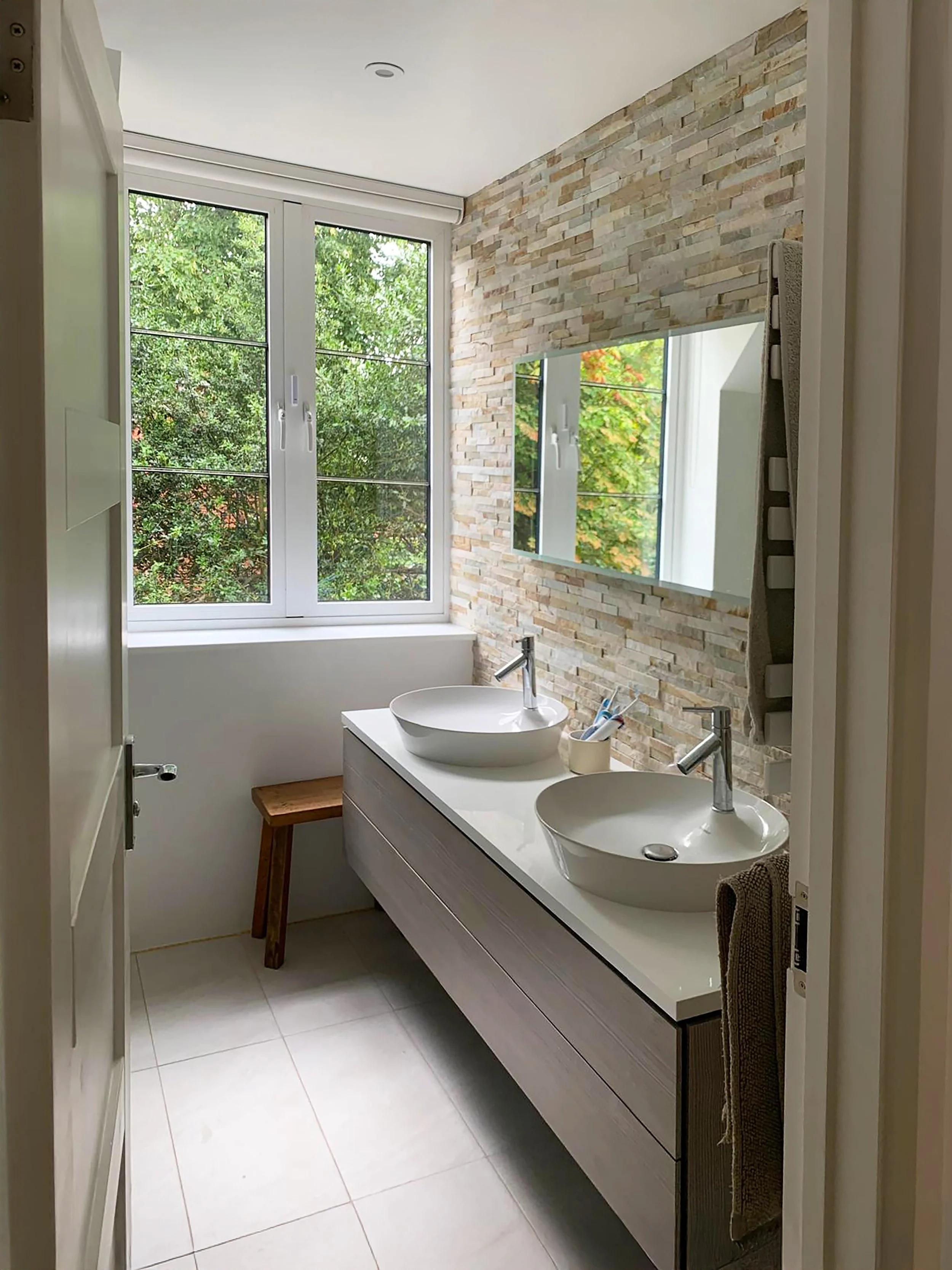 A modern bathroom with a double sink vanity, a large window, a stone accent wall with a rectangular mirror, and a brown towel hanging on a side.