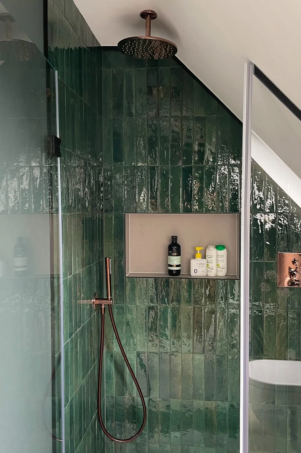 Shower area with green tiles, a rain shower head, a handheld shower hose, and a built-in shelf holding toiletries in a bathroom.