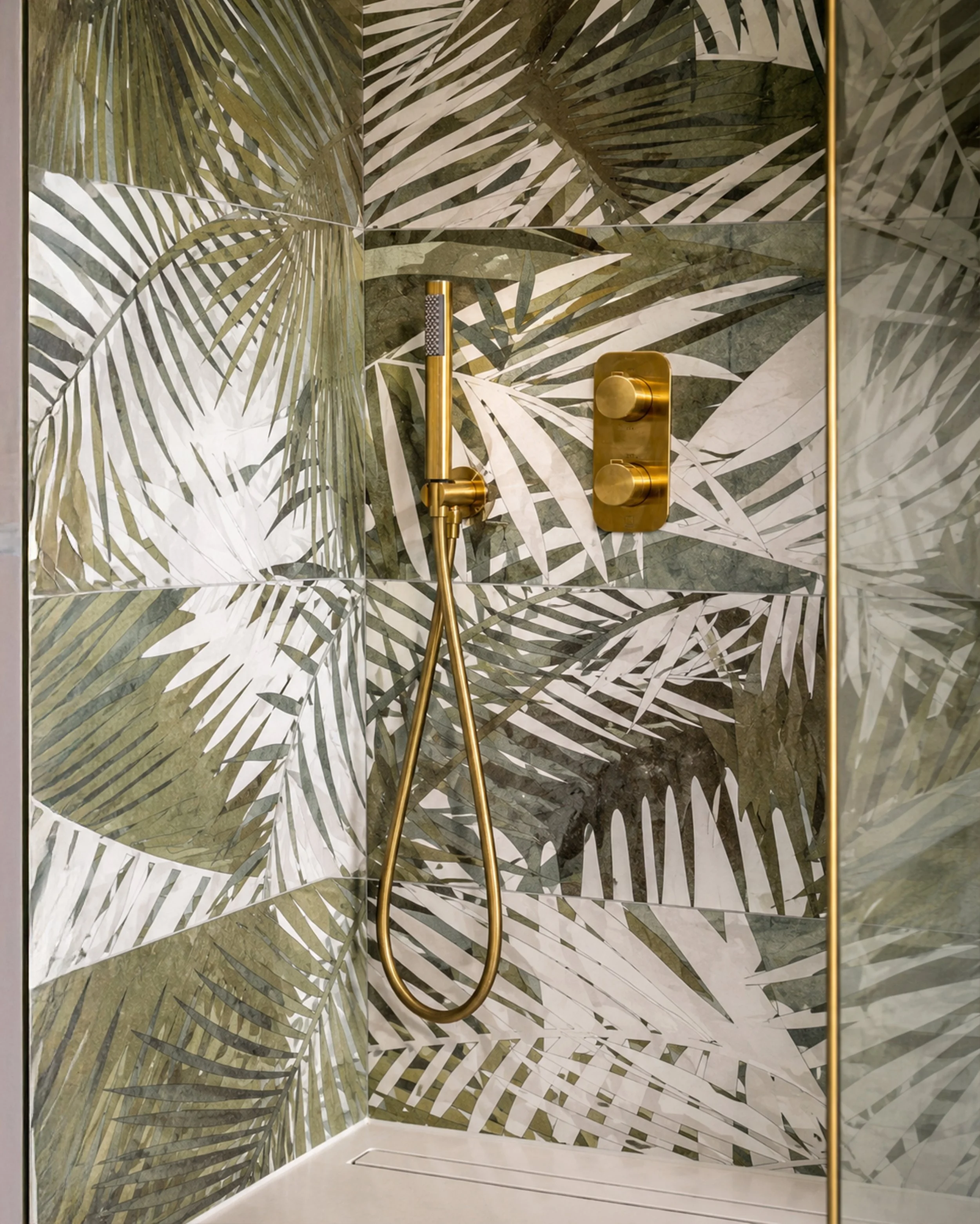 Shower with tropical leaf patterned tiles, gold fixtures, handheld shower head, and glass enclosure